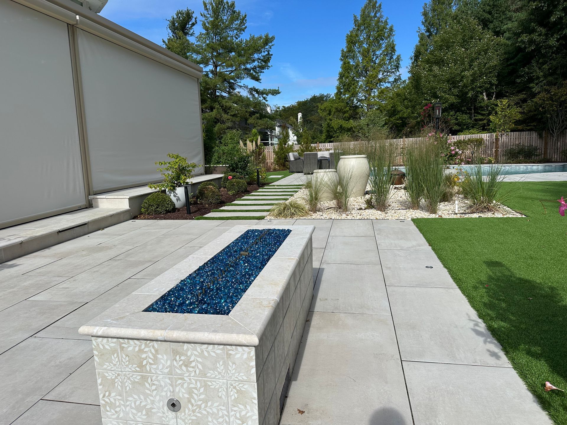 Outdoor patio with a stone fire pit filled with blue glass, surrounded by greenery and stone pavers.
