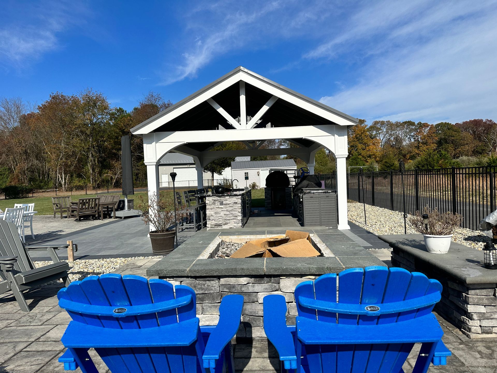 Two blue Adirondack chairs face a stone fire pit under a white gazebo on a patio.