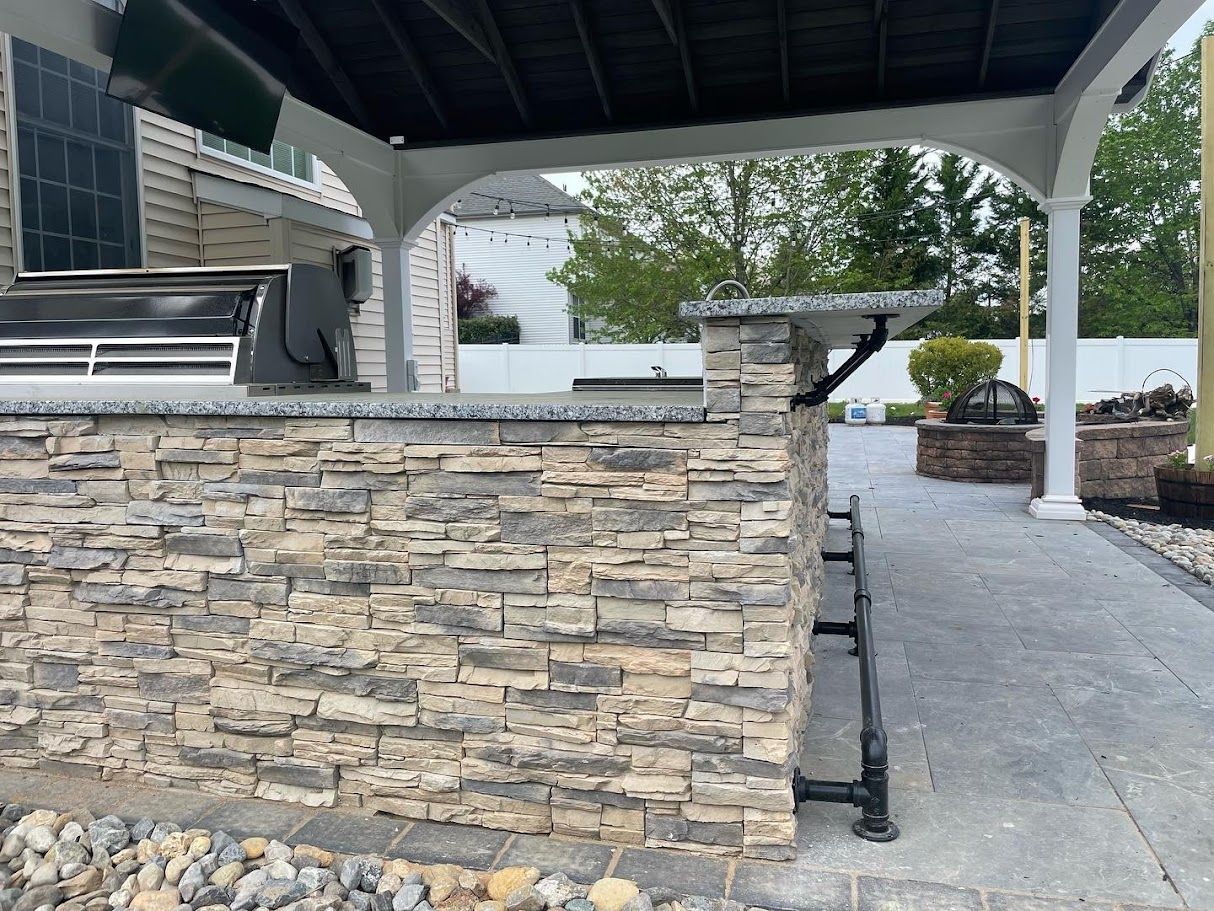 Outdoor kitchen with stacked stone wall, granite countertop, and metal bar seating under a covered patio.