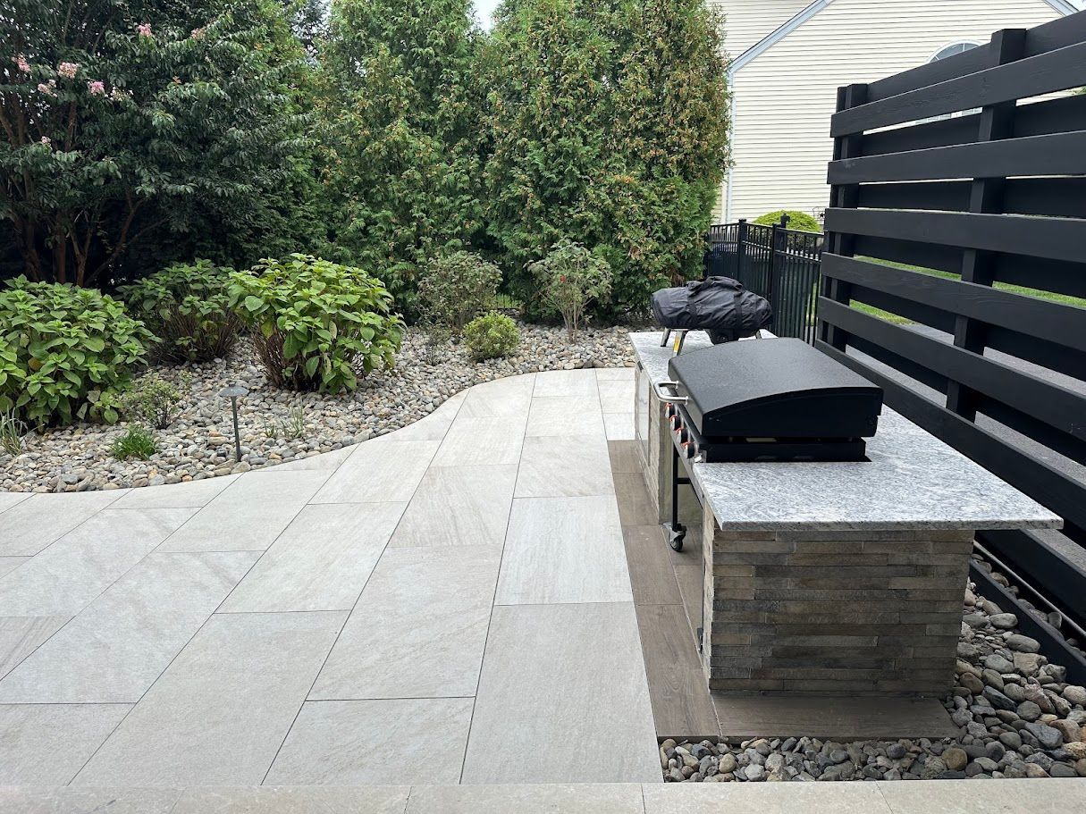 Patio with stone pavers, built-in grill, and landscaping with shrubs and a dark fence.