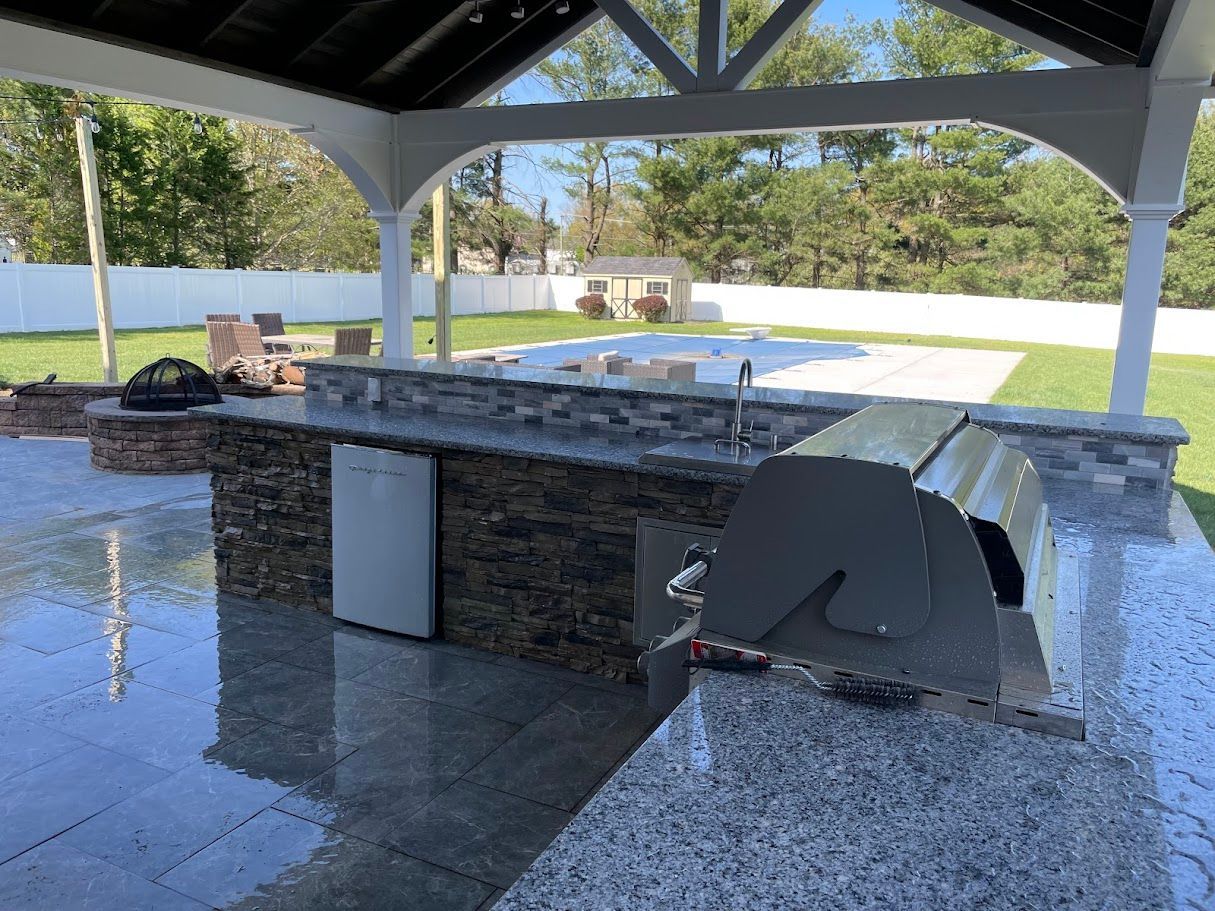 Outdoor kitchen with stone and gray countertops, grill, and under-counter refrigerator under a white gazebo.