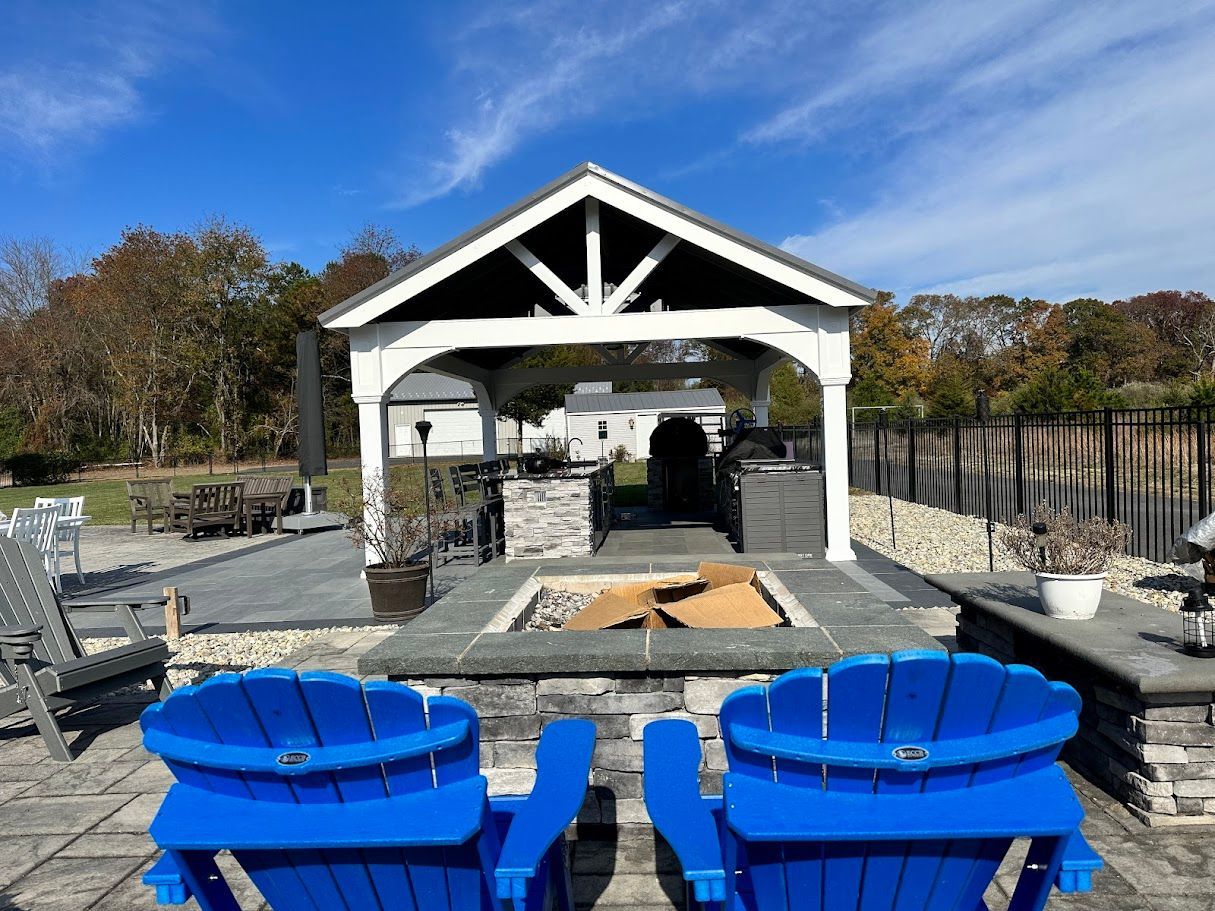 Two blue Adirondack chairs face a stone fire pit under a white gazebo. Sunny outdoor scene.
