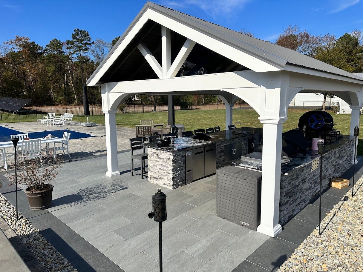 Outdoor kitchen under a white gazebo with gray tile and stone walls, next to a pool.