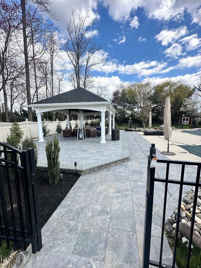 Gazebo on a patio, overlooking a pool area. Blue sky, trees, black fence.