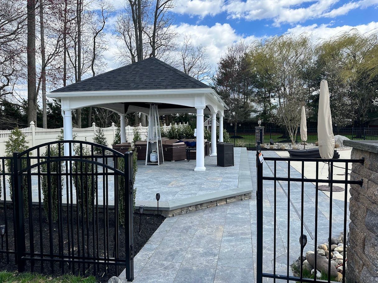 Gazebo with outdoor seating on a paved patio, behind a black metal fence, on a sunny day.