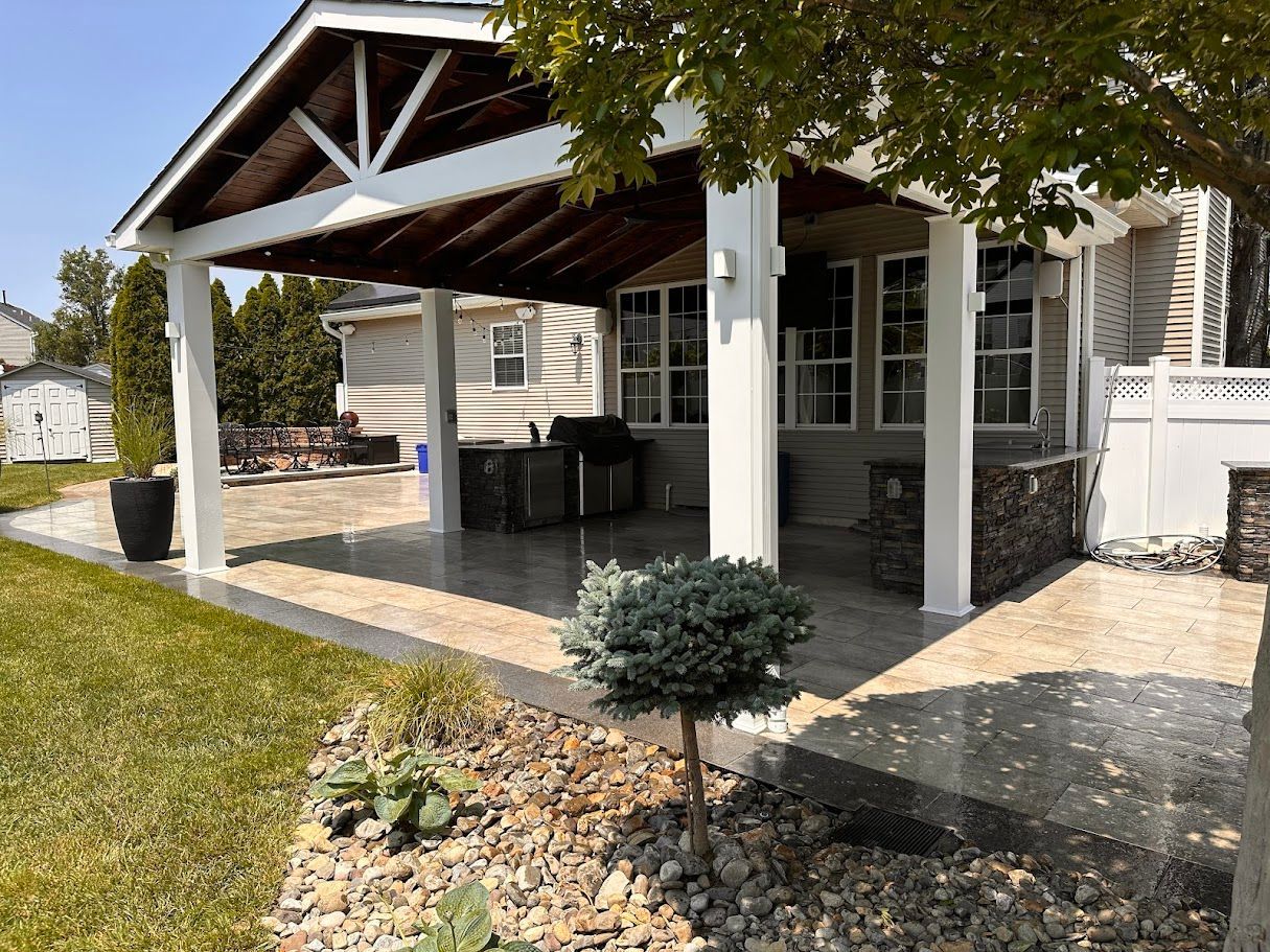 Covered patio with white columns, brown roof, and stone patio with a grassy lawn.