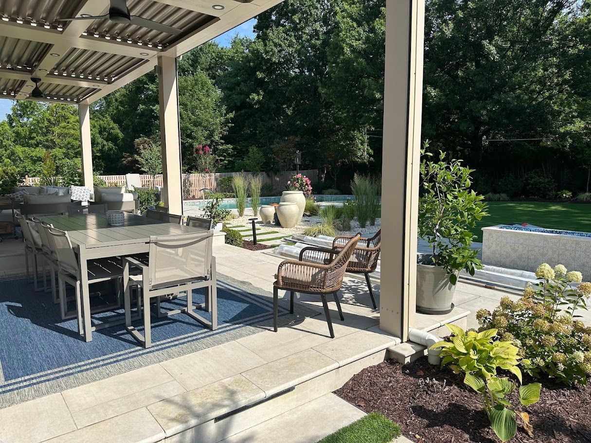 Outdoor patio with dining set, lounge chairs, and landscaping, under a pergola, with a pool visible in the background.