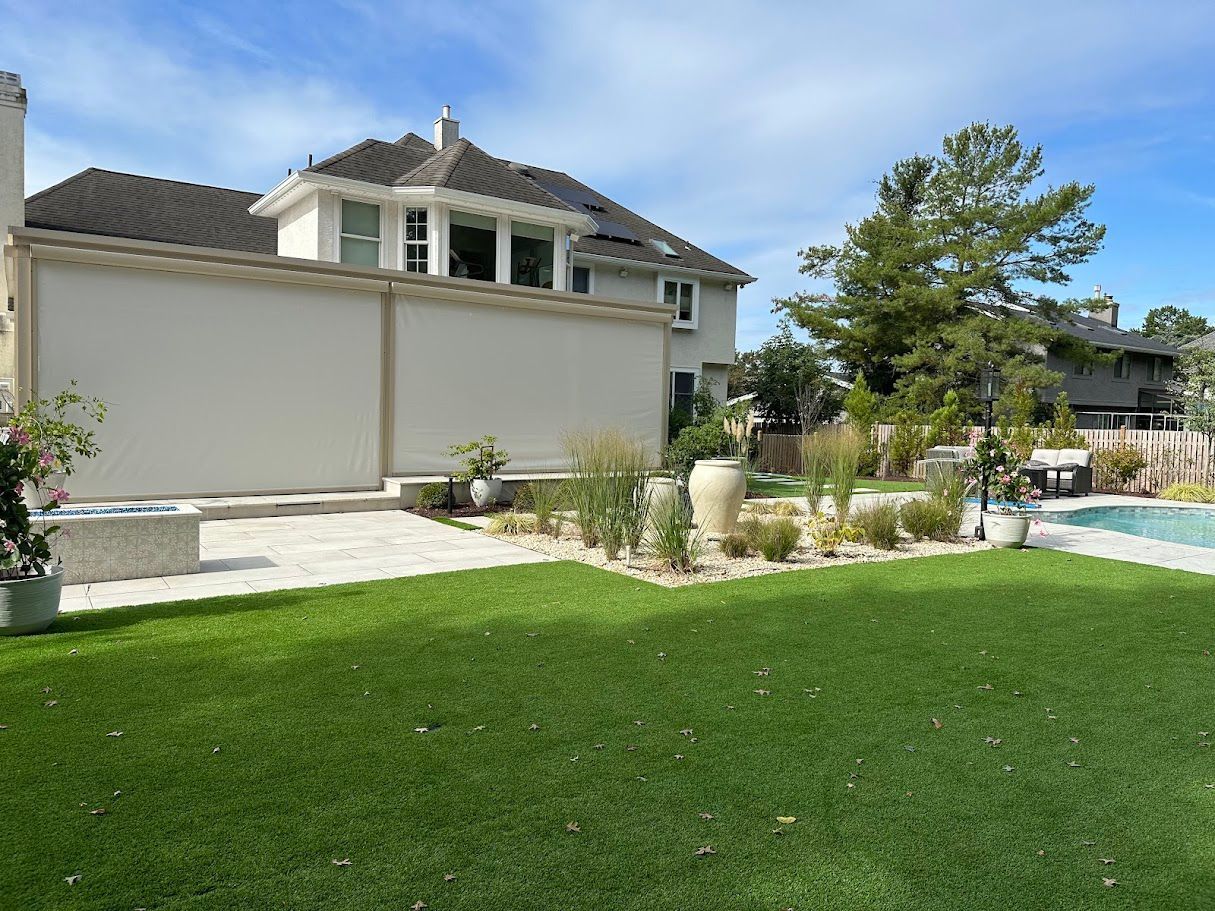Backyard with green lawn, stone patio, beige house, pool, and plants.