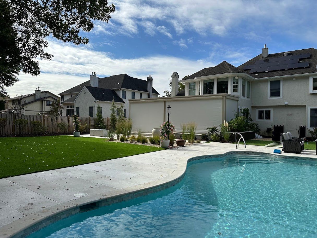 Backyard with a pool, green lawn, and beige house under a blue sky with clouds.