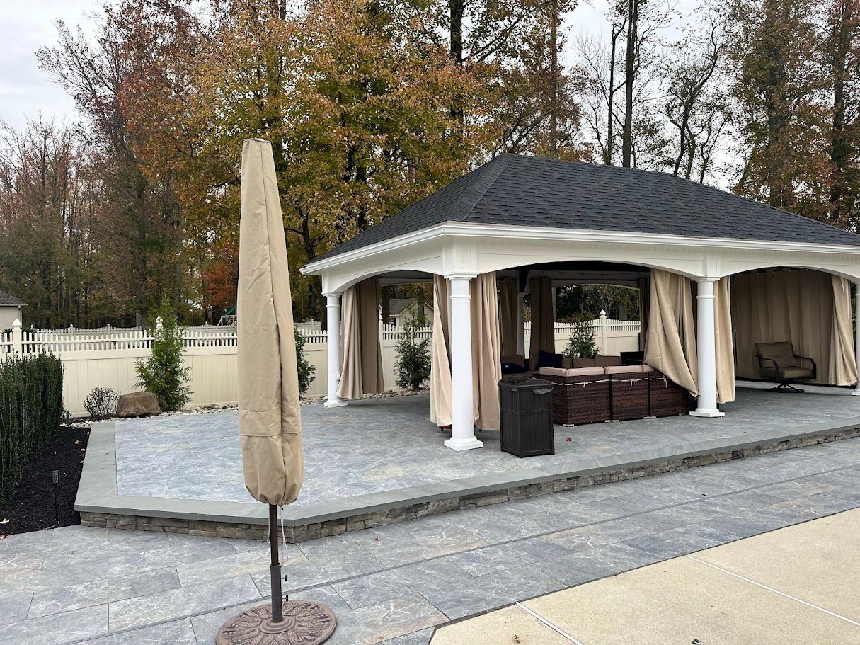Gazebo with beige curtains and black roof on a gray stone patio, umbrella closed.
