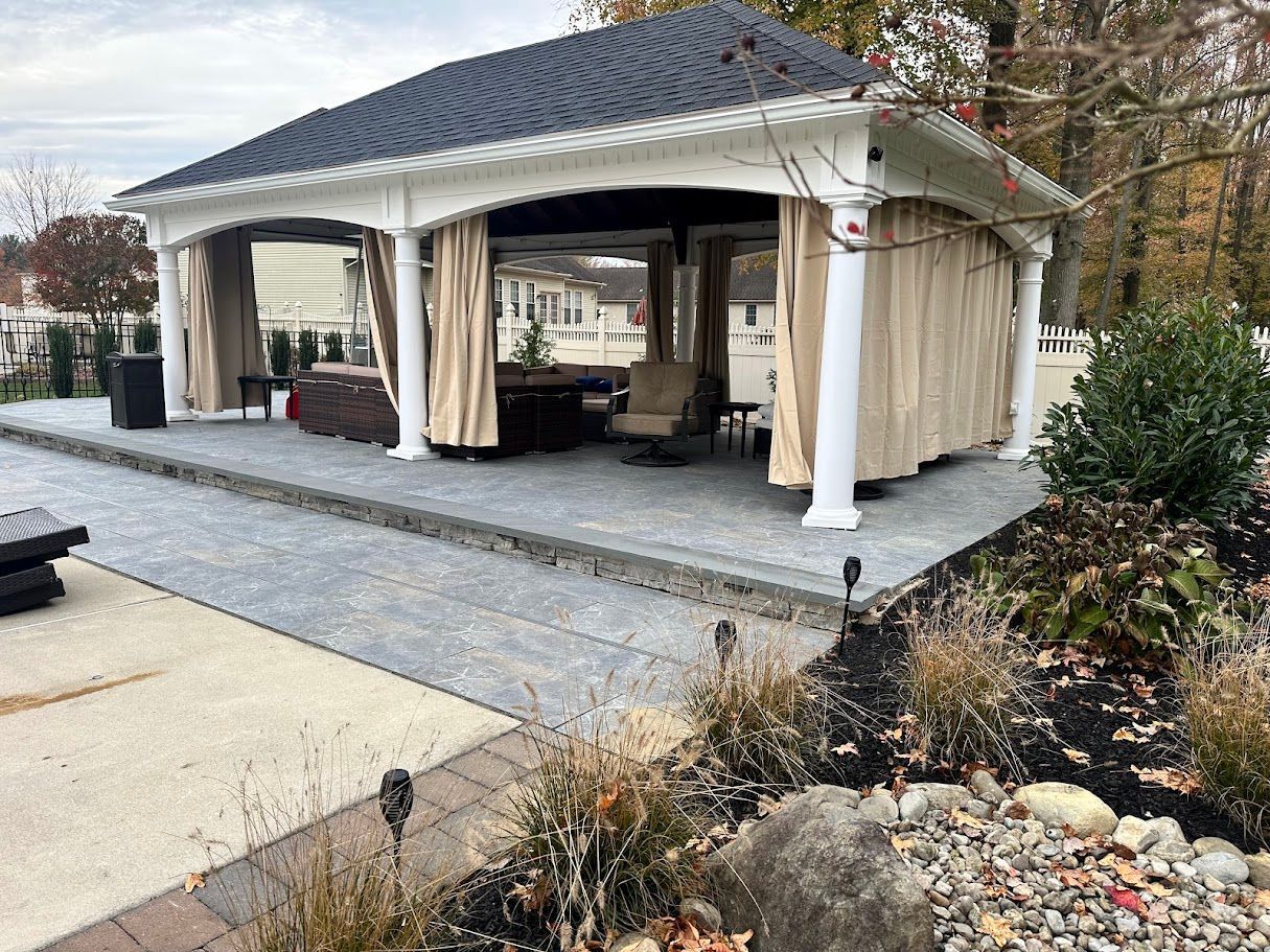 Gazebo with gray stone flooring, tan curtains, white columns. Outdoor seating area.