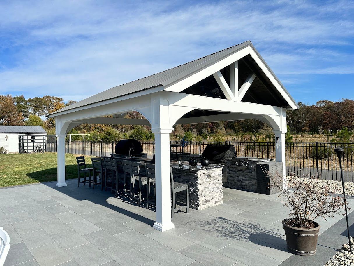 Outdoor kitchen with white pergola, stone base, and grill. Blue sky in the background.