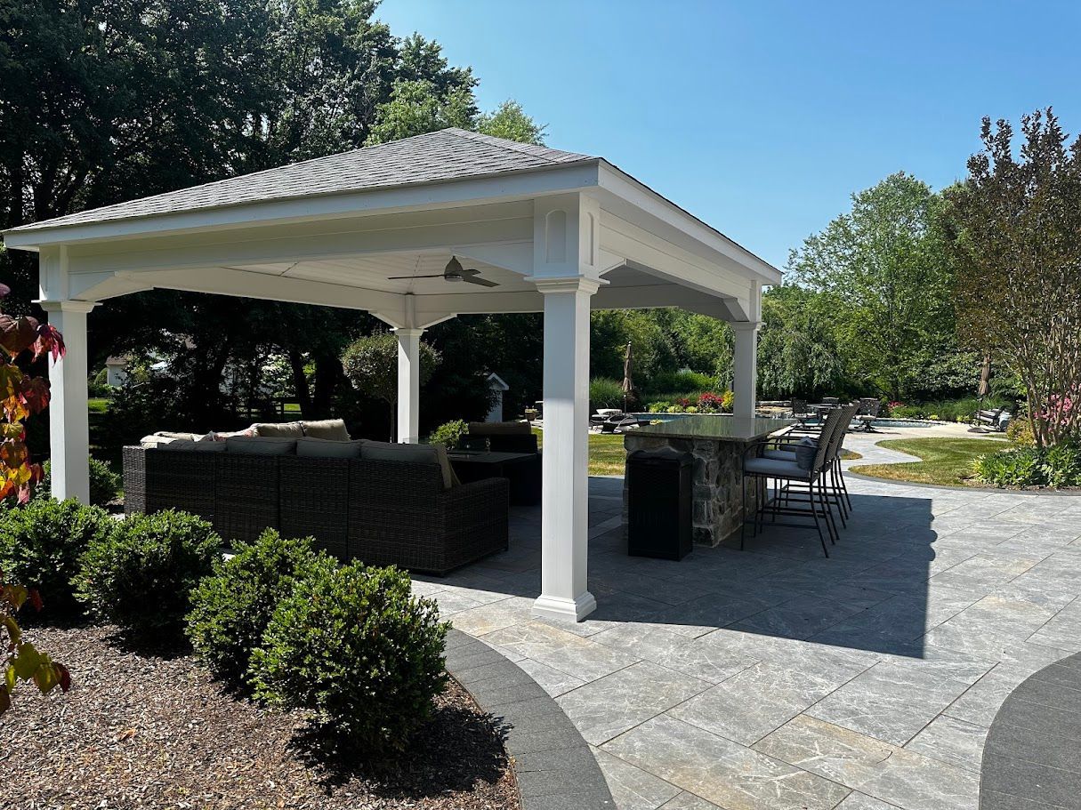 A white gazebo with seating and a bar area on a stone patio in a yard on a sunny day.