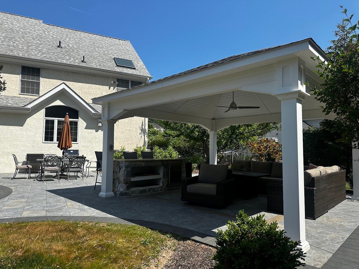 Outdoor patio with seating, grill area, and a white-columned roof, next to a large house.
