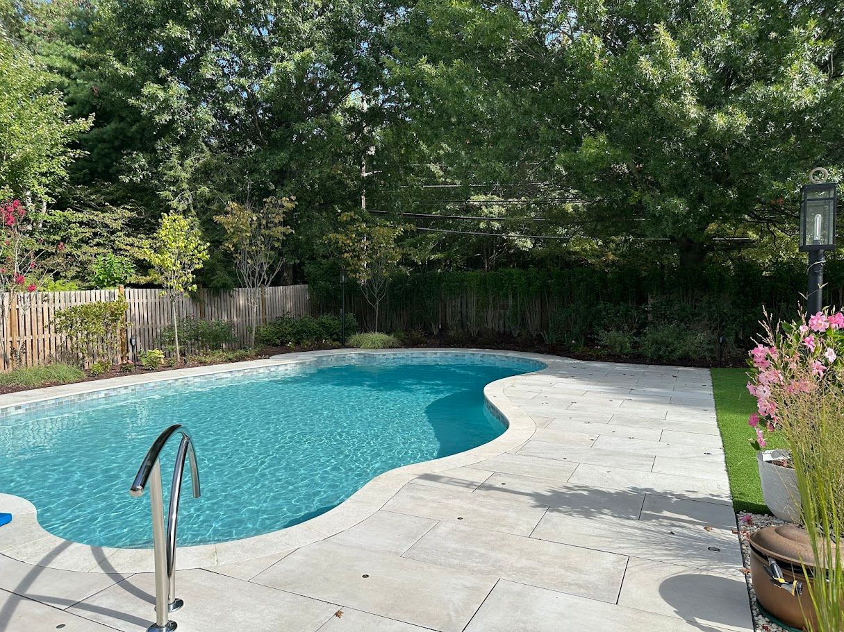 Swimming pool with blue water and stone patio, surrounded by greenery and trees.