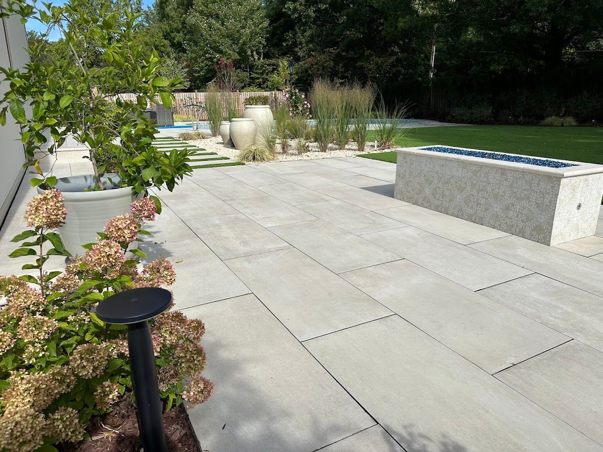 Stone patio with plantings, built-in fire pit, and a manicured lawn. Sunny outdoor setting.