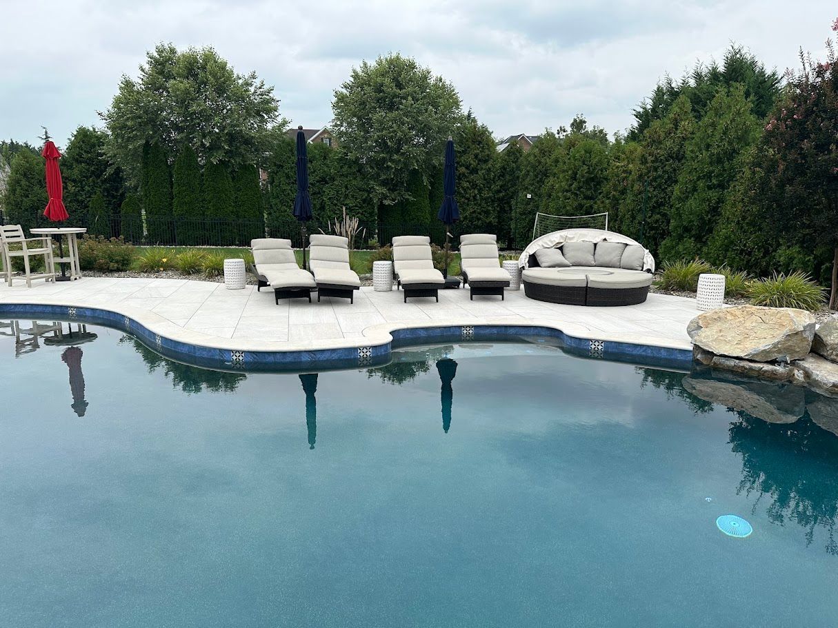 Poolside scene with lounge chairs, umbrella, and a round sofa, with trees and a cloudy sky in the background.