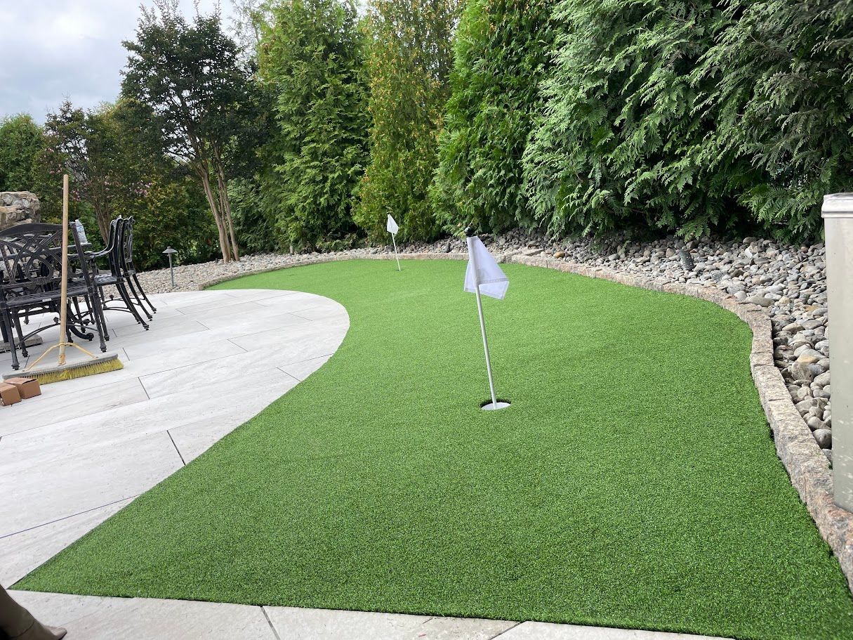 Green artificial turf putting green with a white flag, bordered by stone and landscaping.