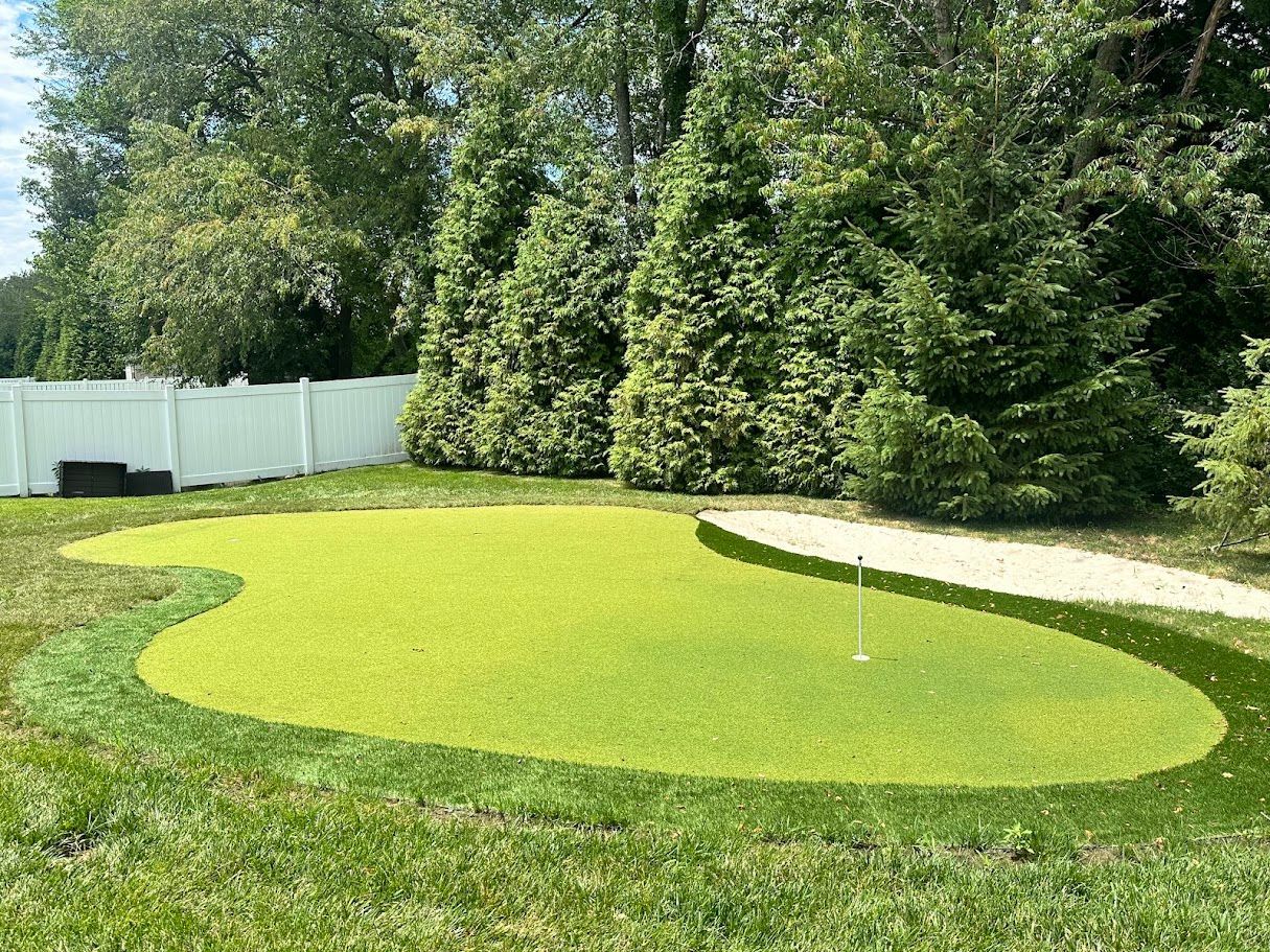 Backyard putting green with sand trap, surrounded by grass and trees, against a white fence.