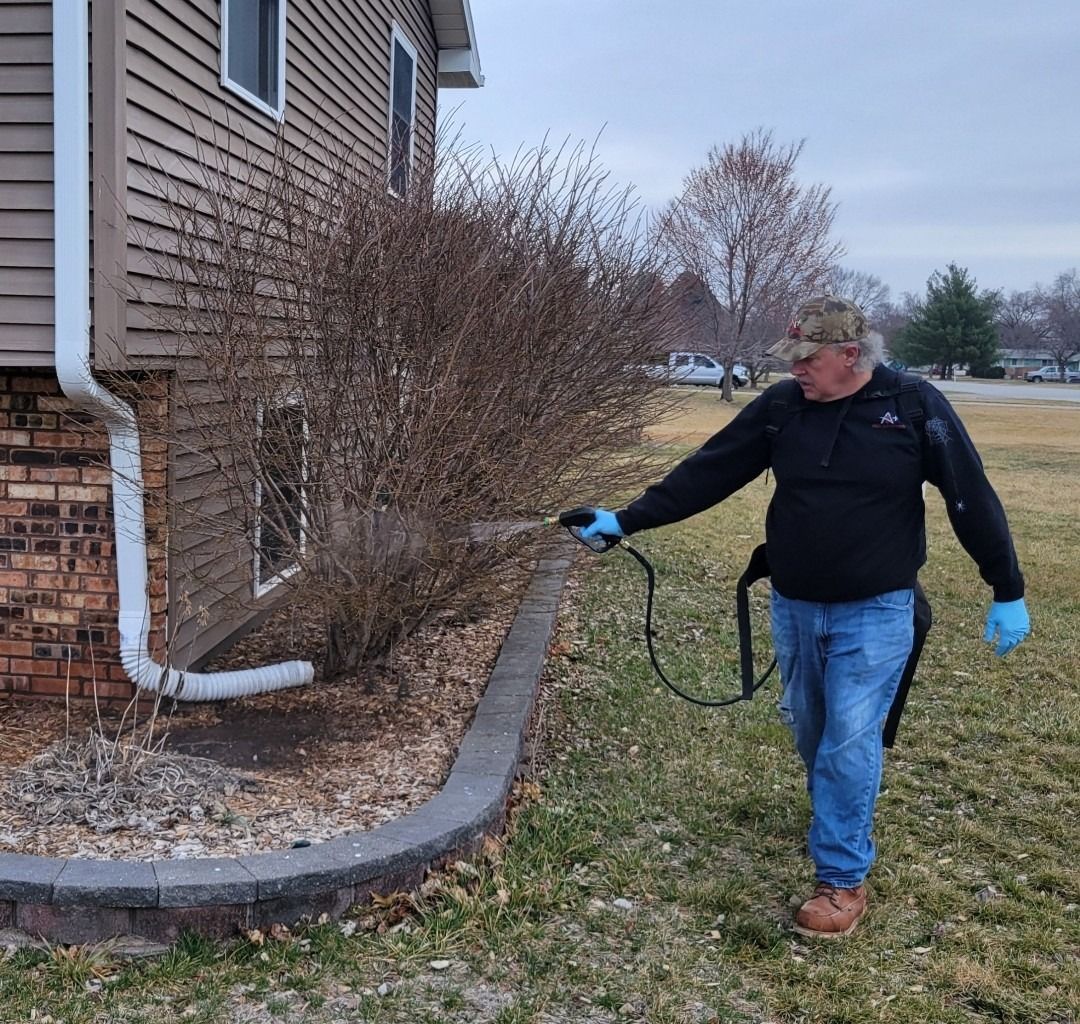 Man spraying bushes next to a building. Brown building with a white downspout and stone border in a grassy setting.