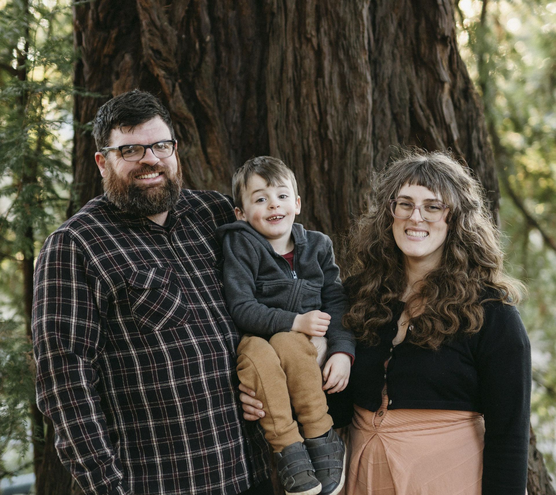 Family of three poses in front of a large tree. Man holds a child, woman stands next to them.