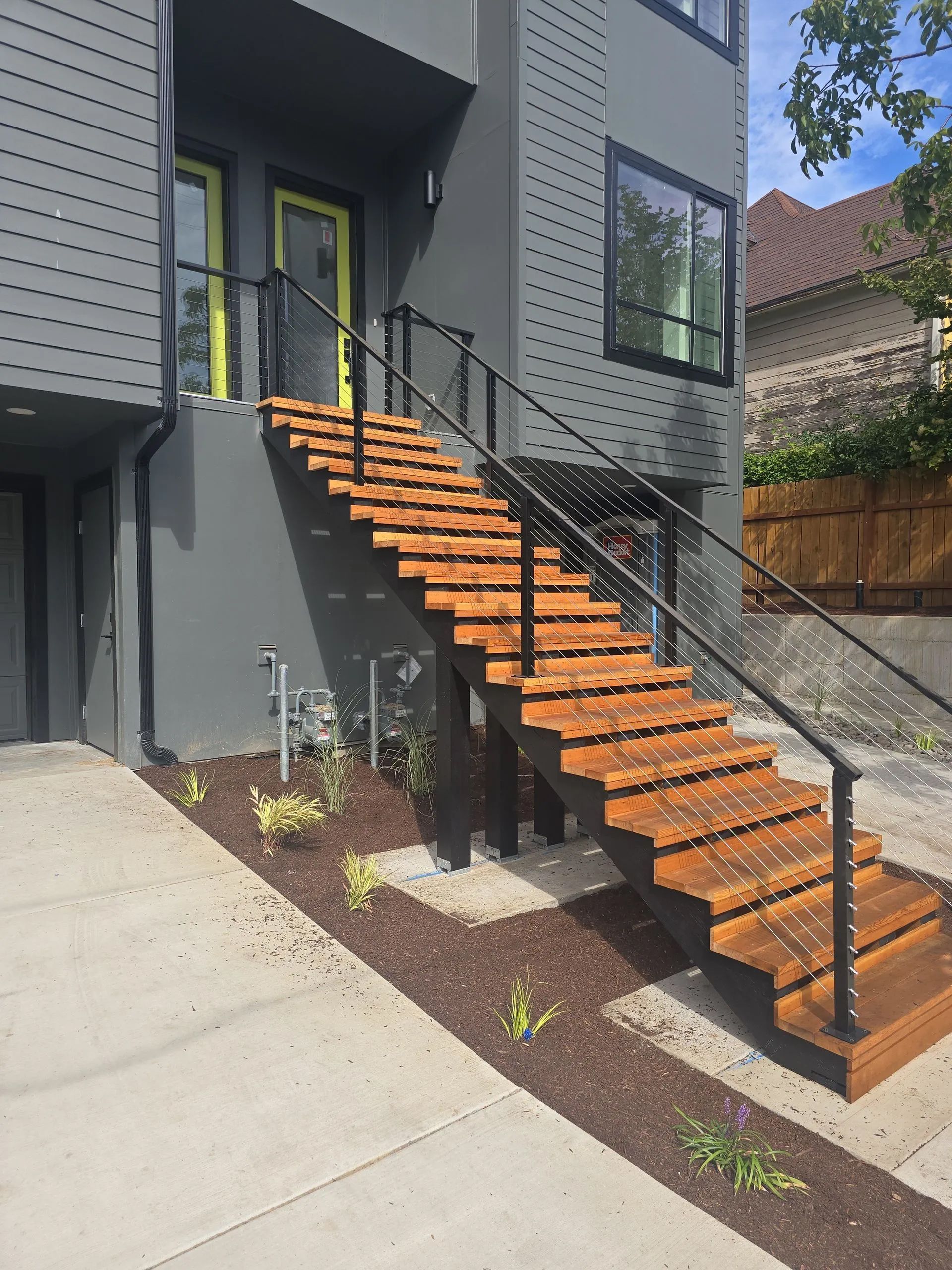 Exterior stairs lead to a modern building with gray siding, a green door, and black handrails.