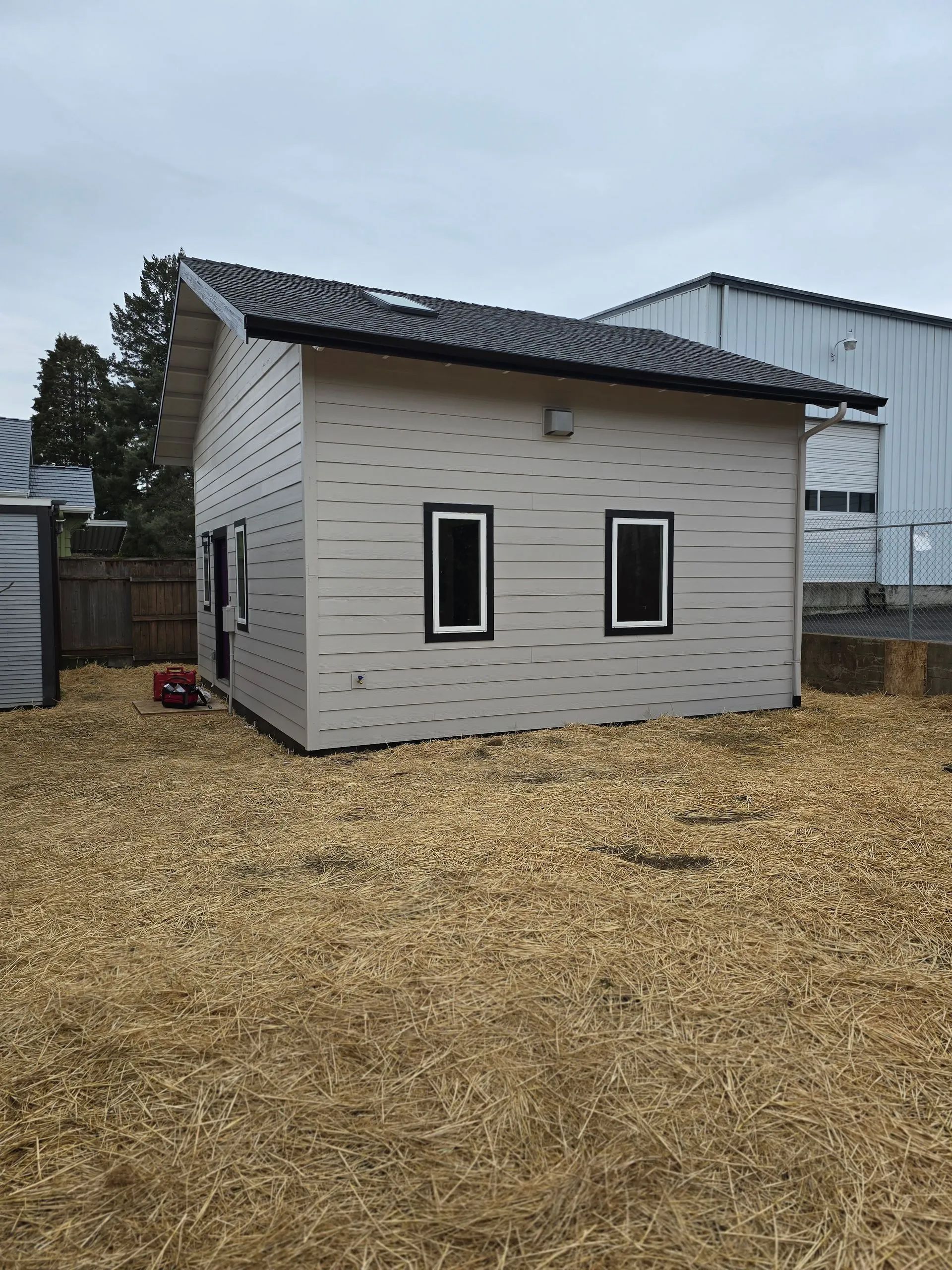 Small, light-colored building with two rectangular windows and a dark roof. Located in a grassy area under an overcast sky.