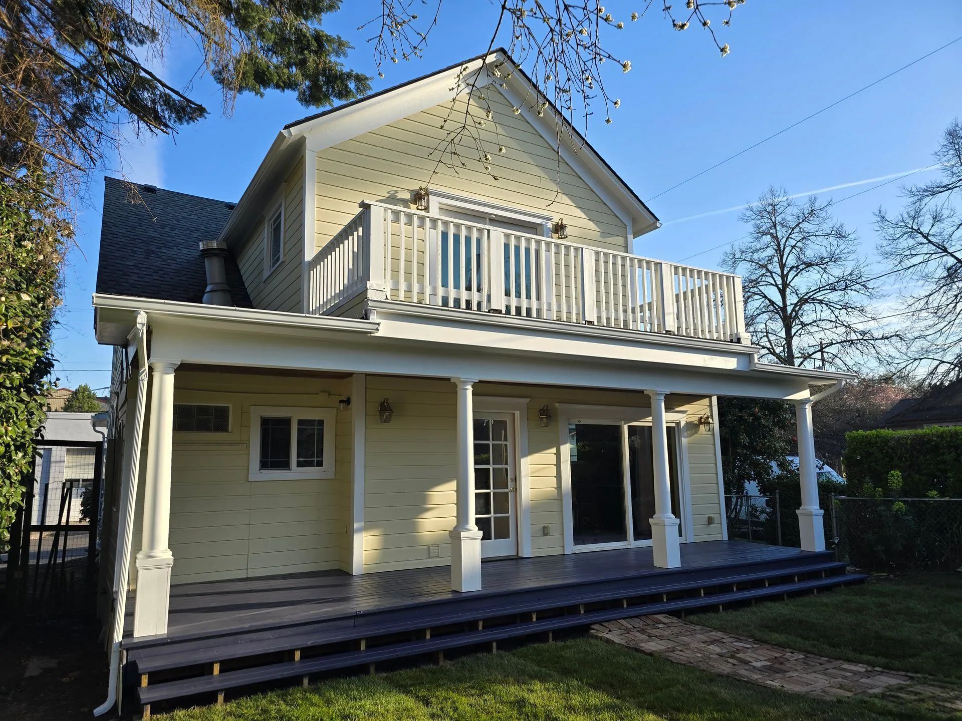 Yellow two-story house with porch and balcony. Blue sky, green grass. White trim, dark blue deck.