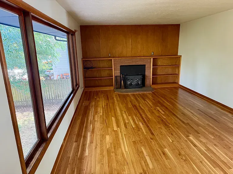 Empty living room with wood flooring, large windows, fireplace, and built-in shelves.