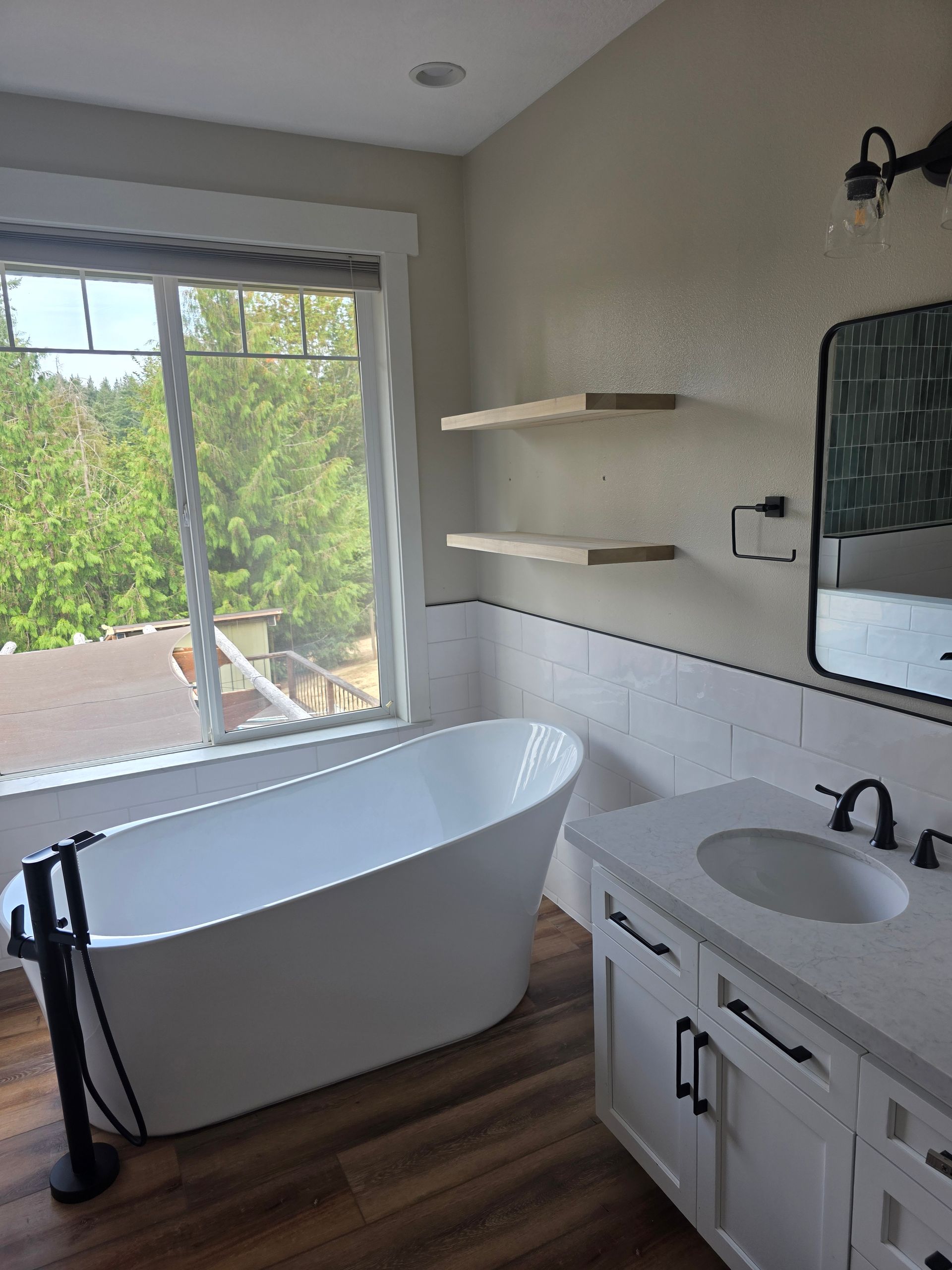Bathroom with white tub, vanity, and shelves; large window with trees outside.