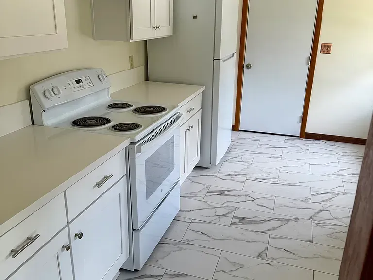 White kitchen with electric stove, cabinets, refrigerator, and marble-look tile flooring.