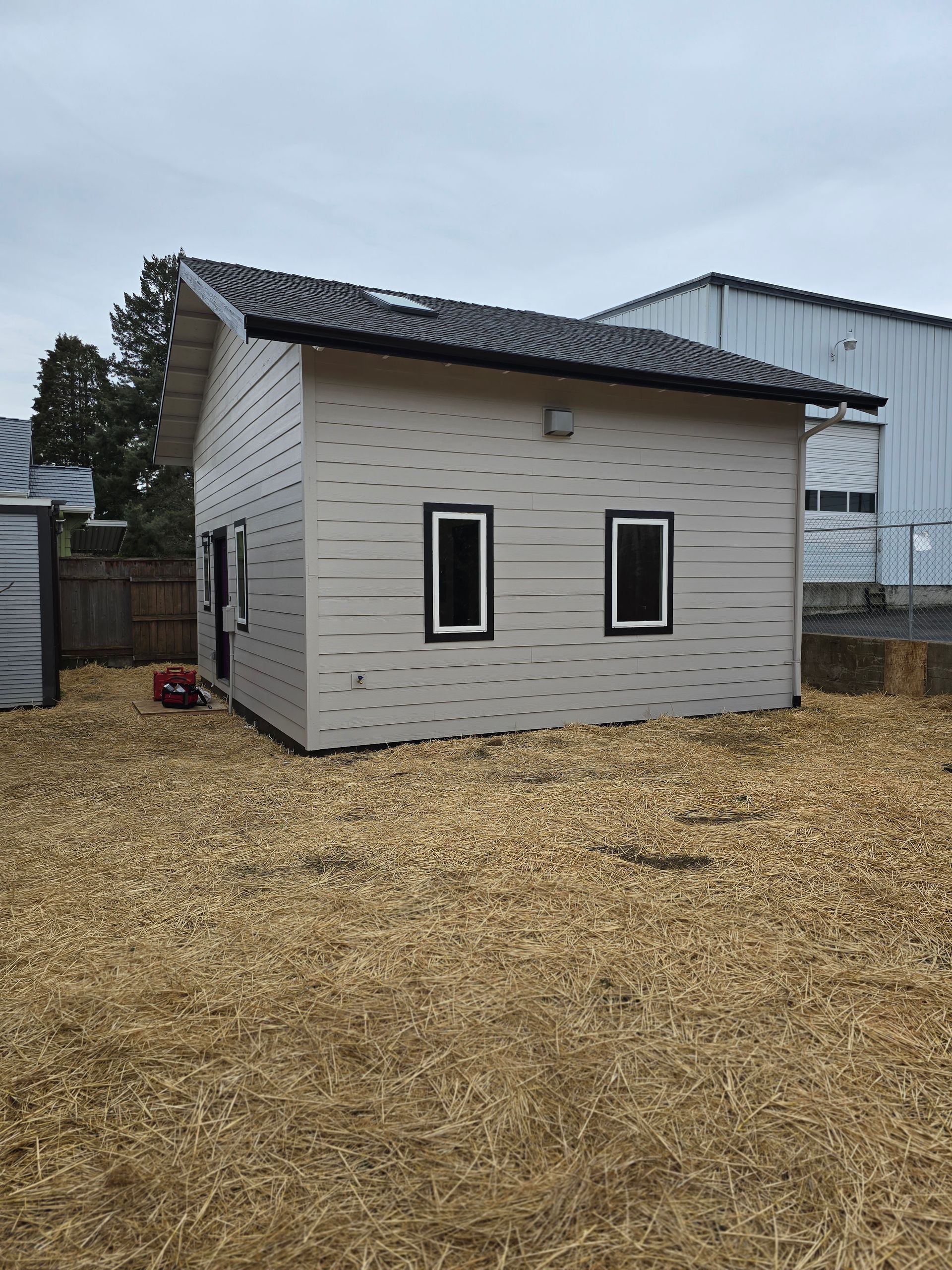 Small, light-colored building with two black-framed windows and a dark roof, set on a bed of wood chips.