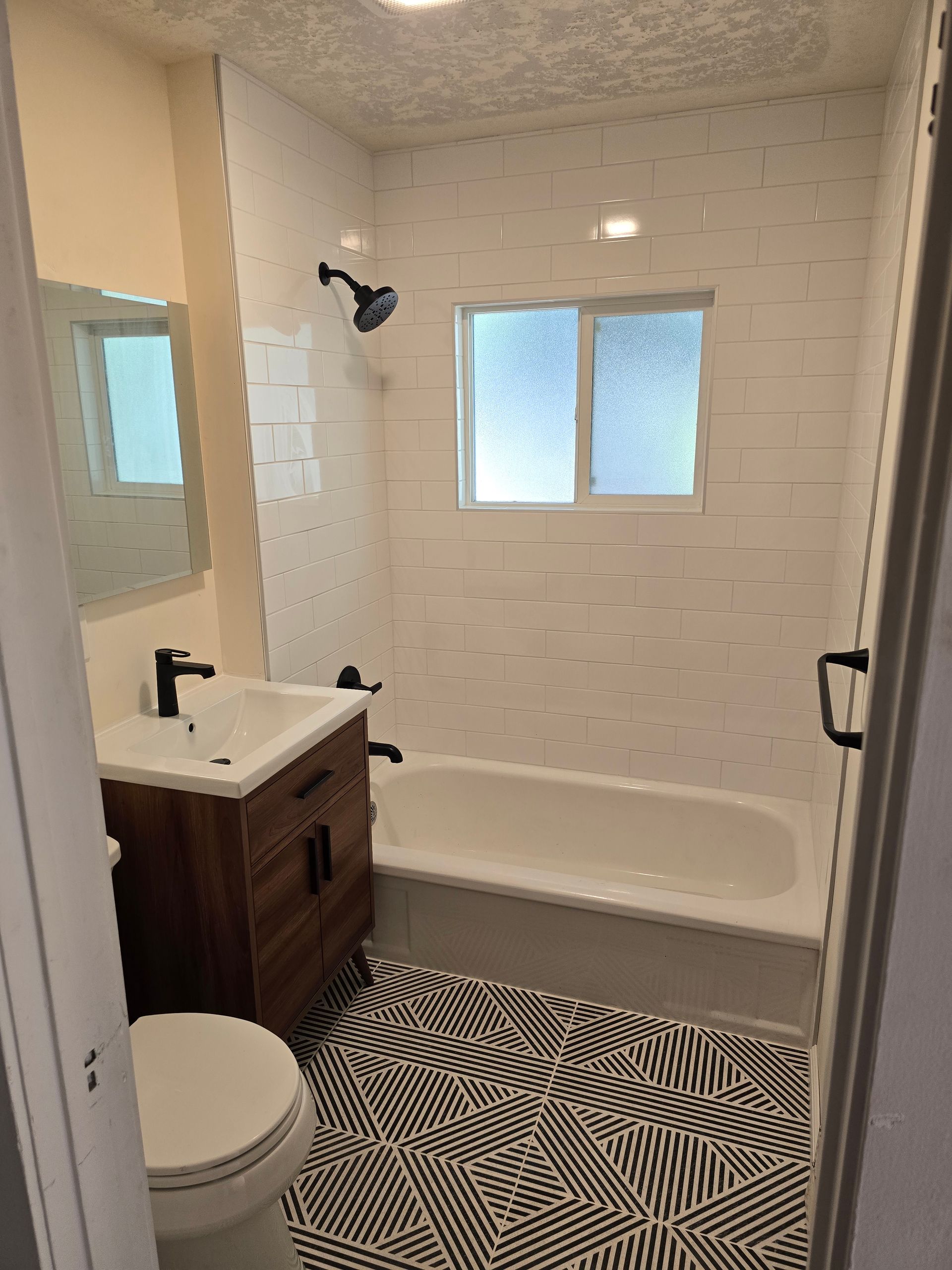 Bathroom with black and white patterned floor, white tiled walls, dark fixtures, and wooden vanity.