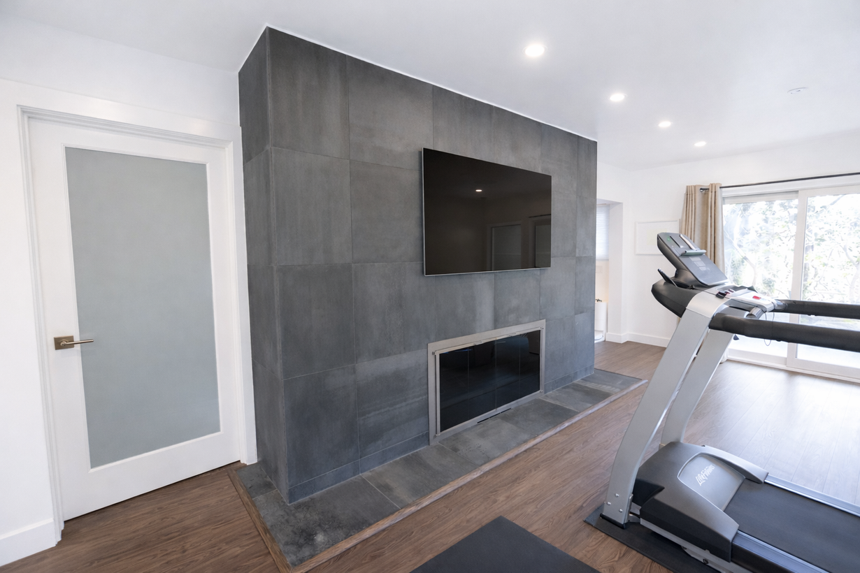 A fitness room with a treadmill facing a large, dark gray tiled fireplace wall with a mounted television.