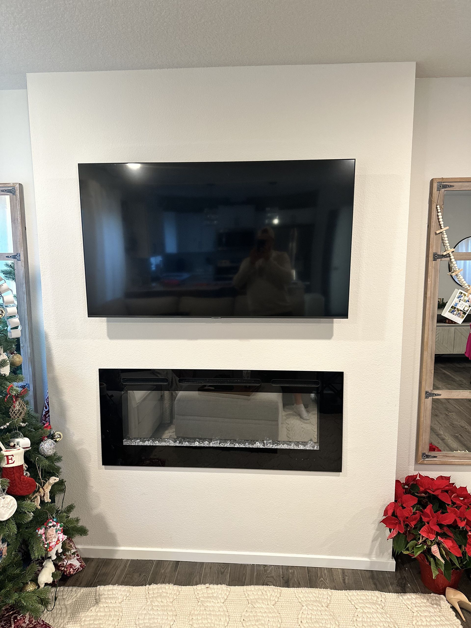 Modern living room wall with mounted TV above a black electric fireplace, flanked by holiday decorations and a mirror.