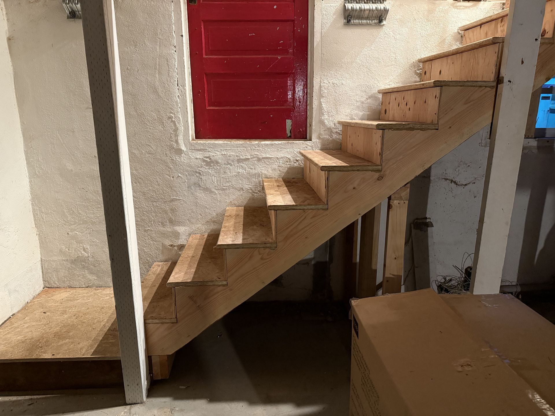 Wooden staircase in a small indoor room with a red door and boxes nearby