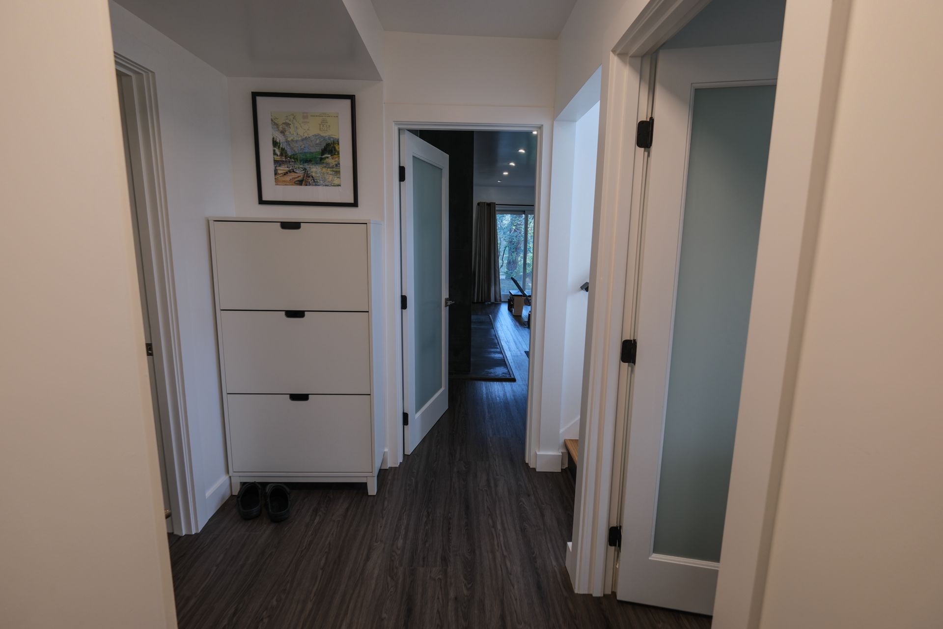 Narrow hallway with white doors, a framed picture, and a white cabinet at the end, leading to a dark room