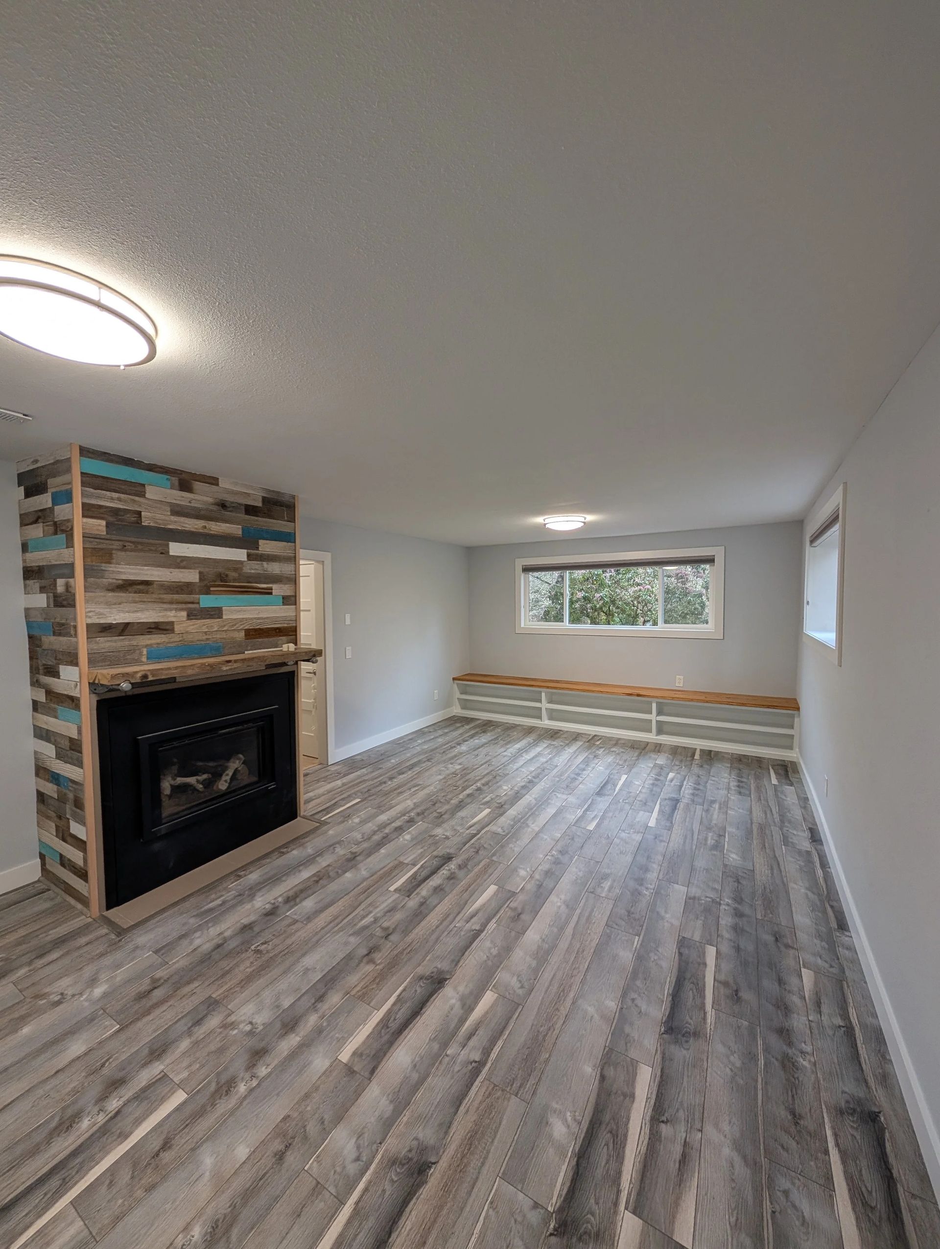 Empty living room with wood-look floors, brick fireplace, and a window bench under a large window.
