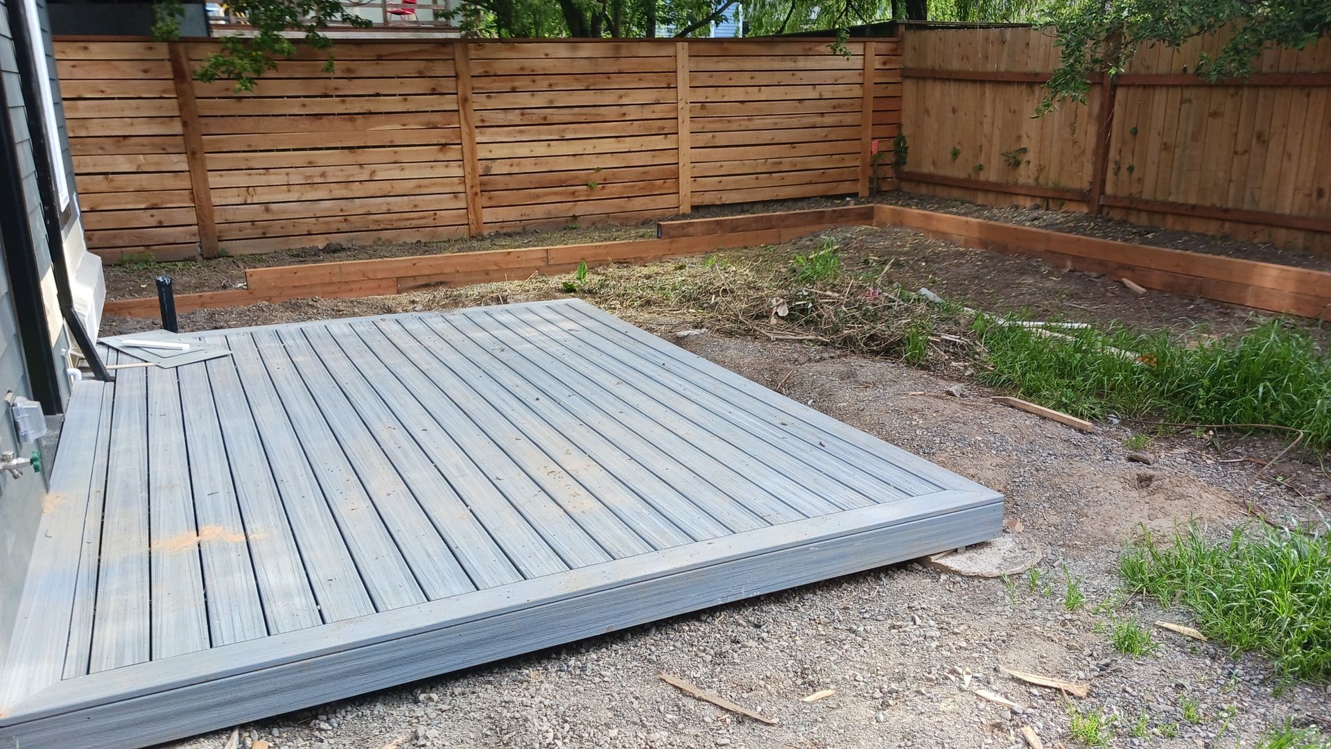 Gray wooden deck platform in a fenced backyard beside a gravel and dirt area