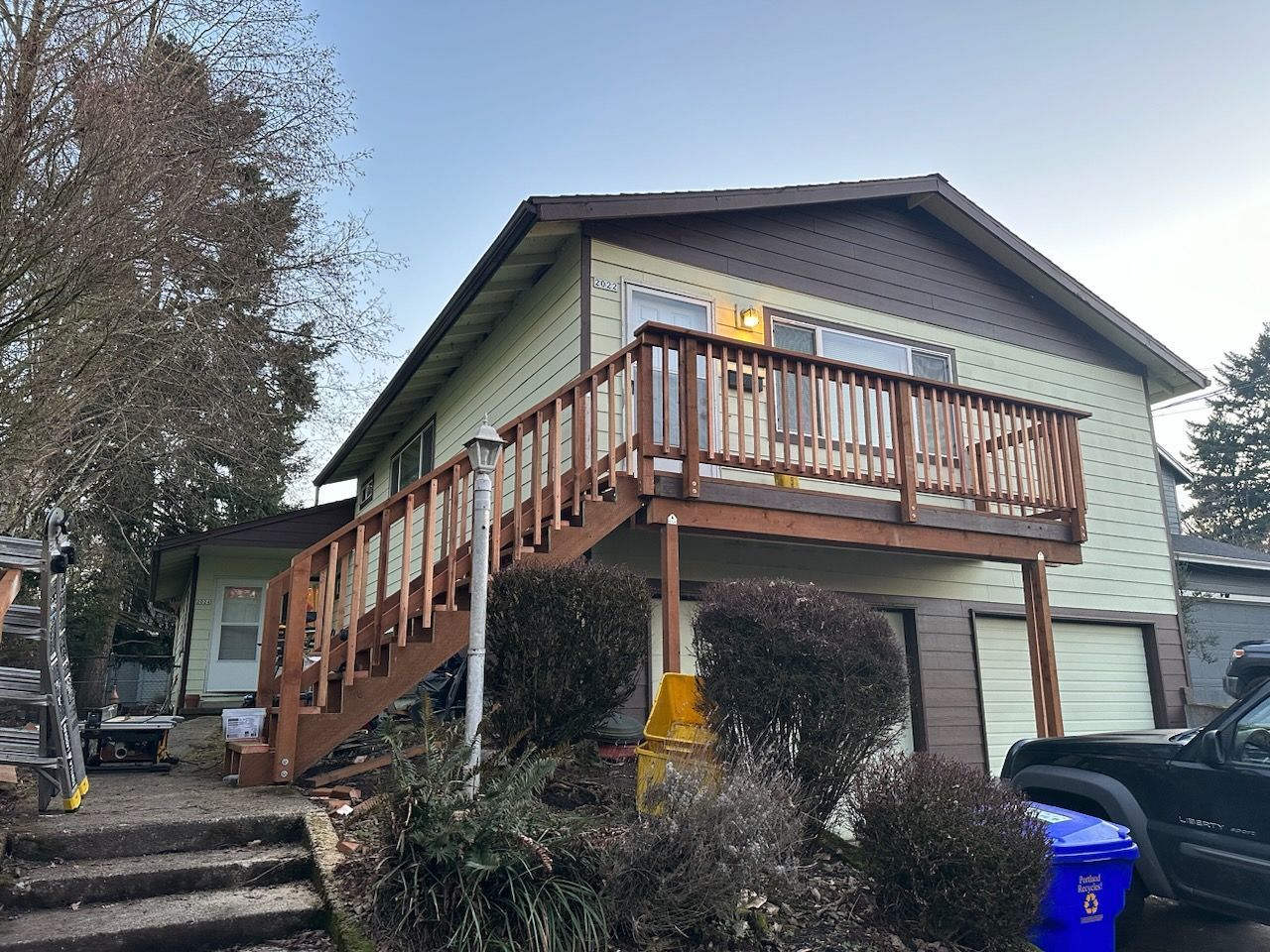 Raised house with exterior wooden stairs, front deck, and blue recycling bin in a residential yard.