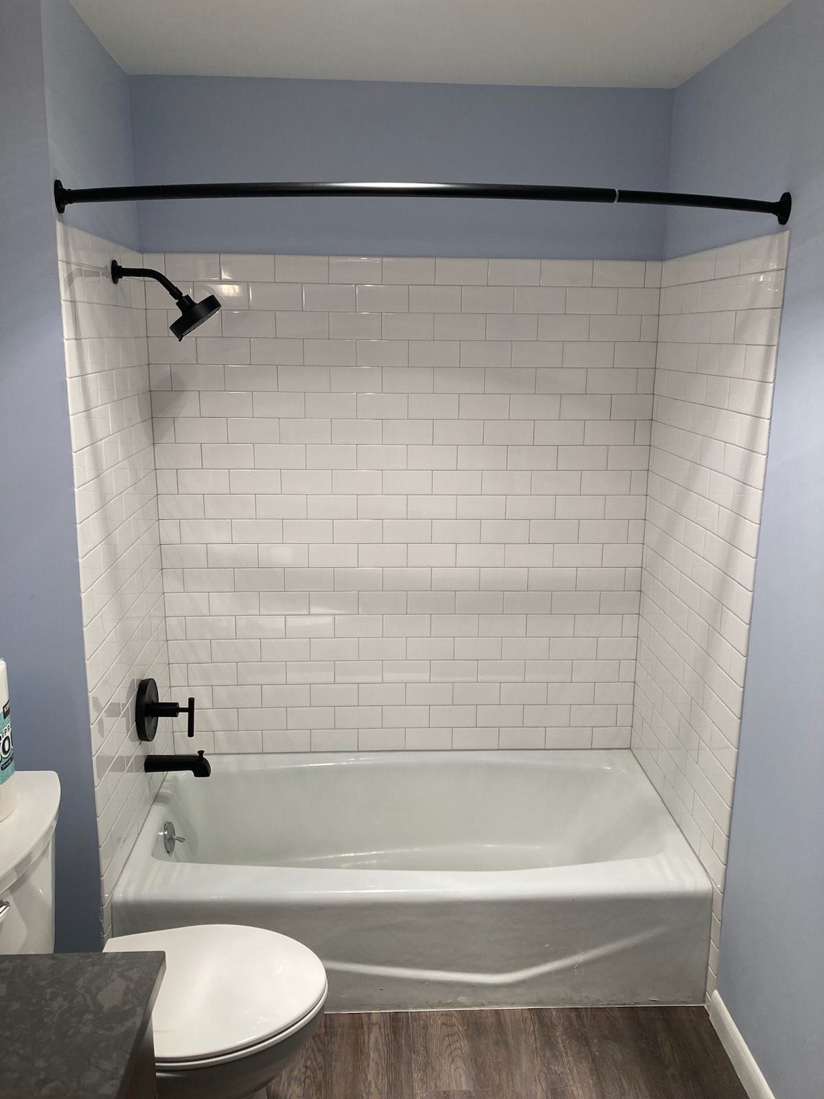 White tiled bathtub and shower alcove with black fixtures, light blue walls, and a toilet in the foreground