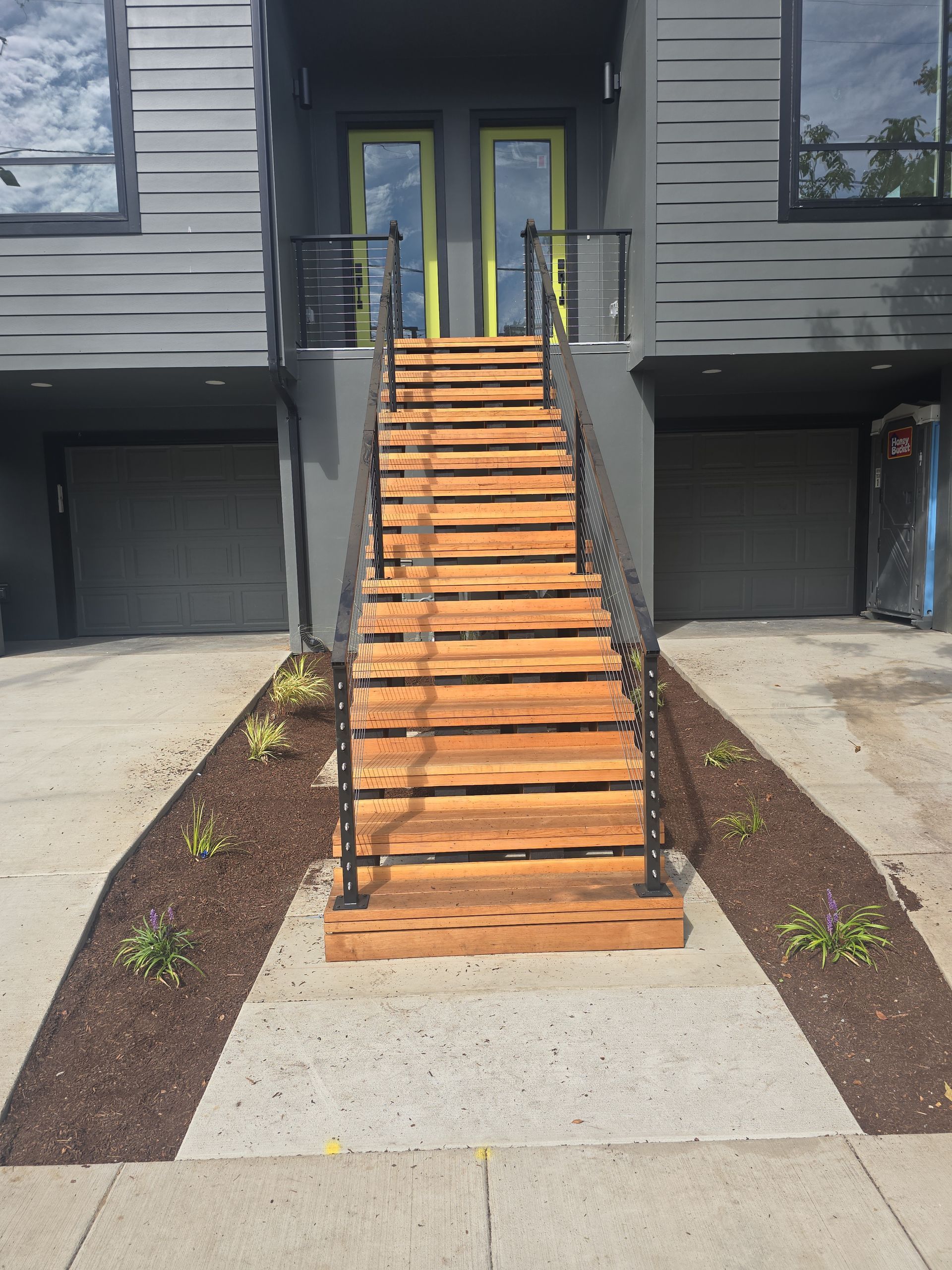 Front entrance with wooden stairs leading to double glass doors, flanked by dark mulch beds and concrete walkways.