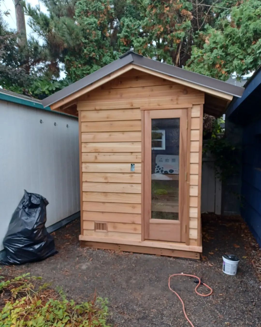 Small wooden shed with a glass door in a narrow backyard between two buildings.
