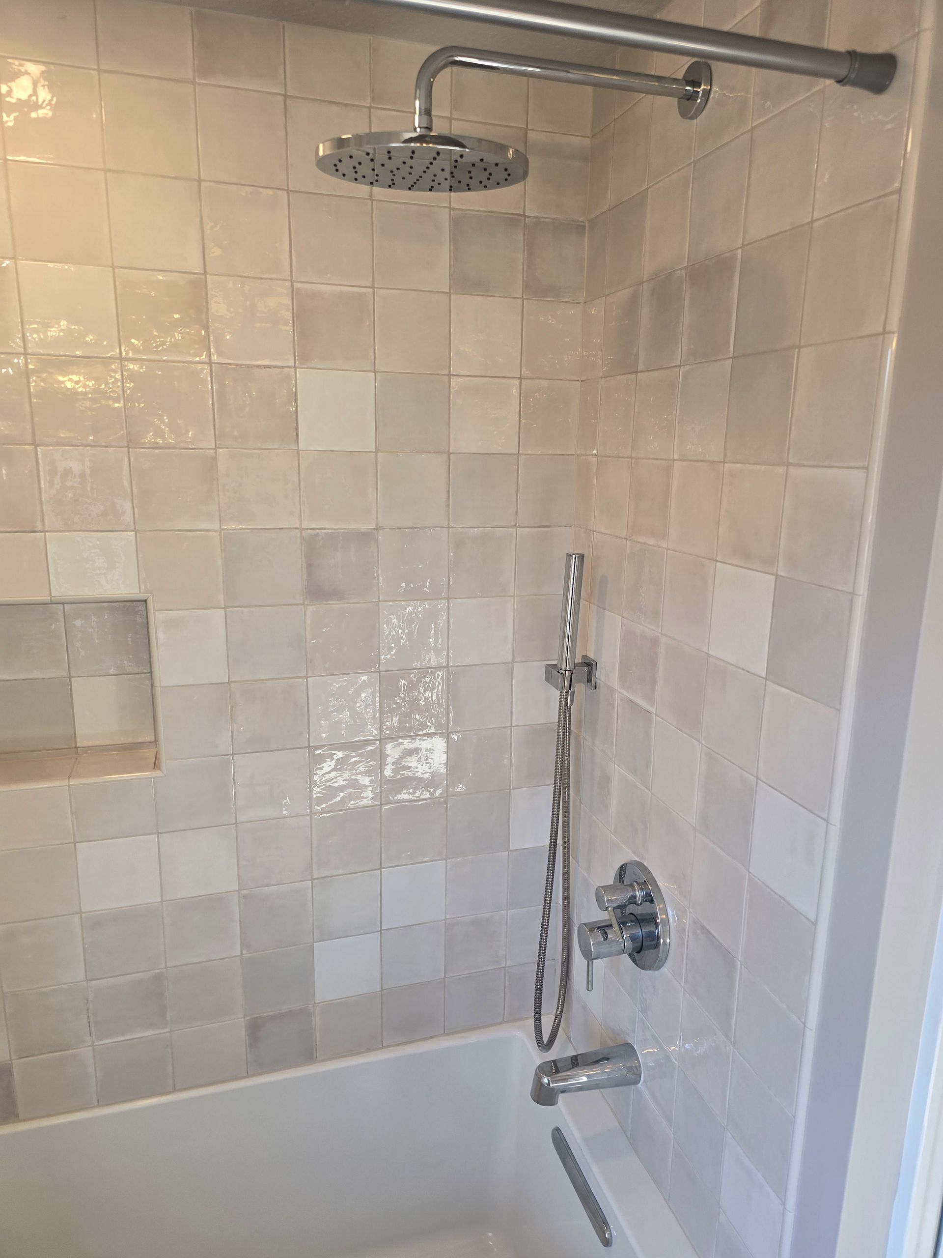 Tiled shower stall with showerhead, handheld sprayer, and wall-mounted controls.