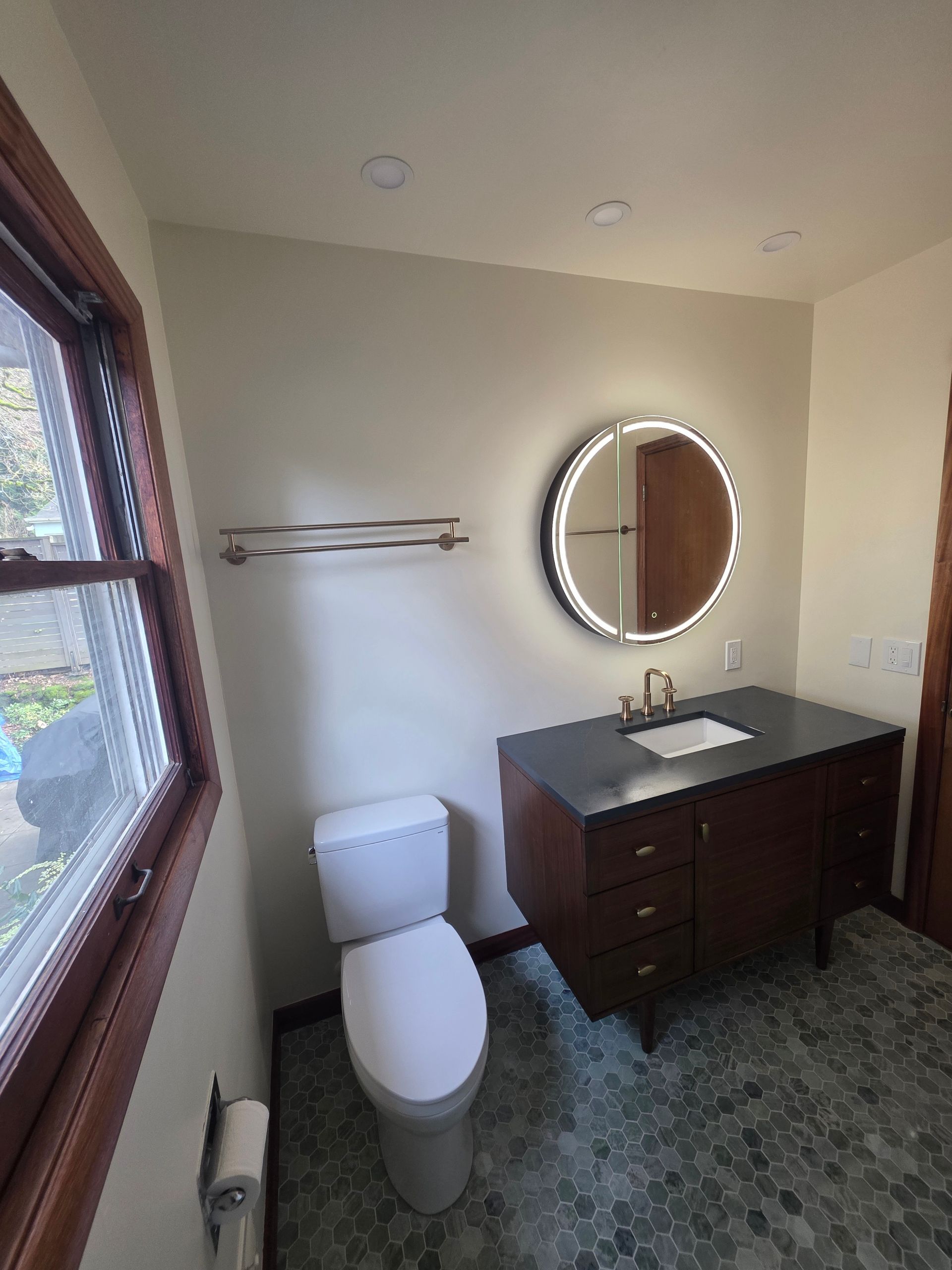 Small bathroom with toilet, dark vanity, round mirror, window, and towel bar.