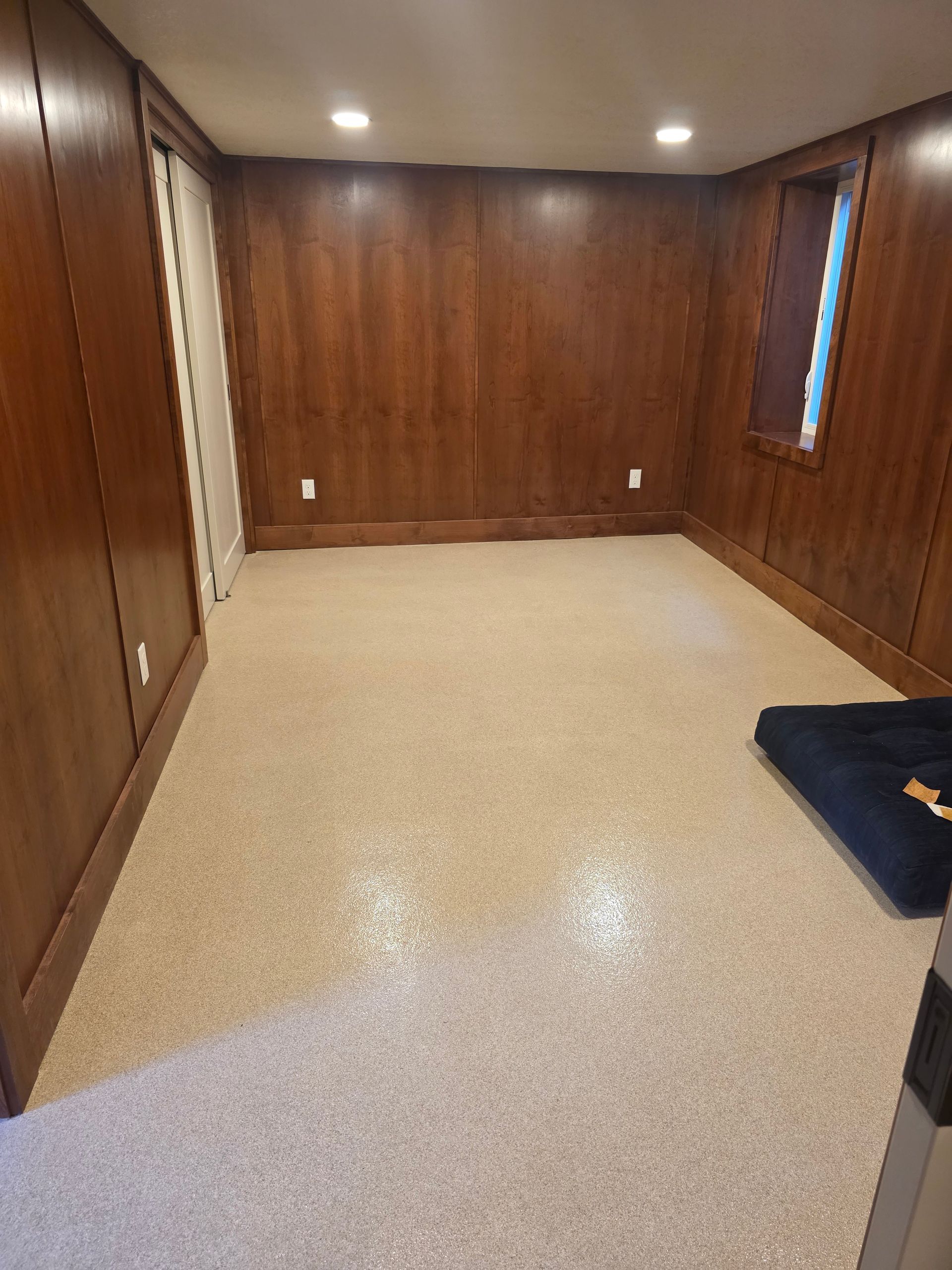 Empty wood-paneled room with beige carpet, recessed lights, and a small window.