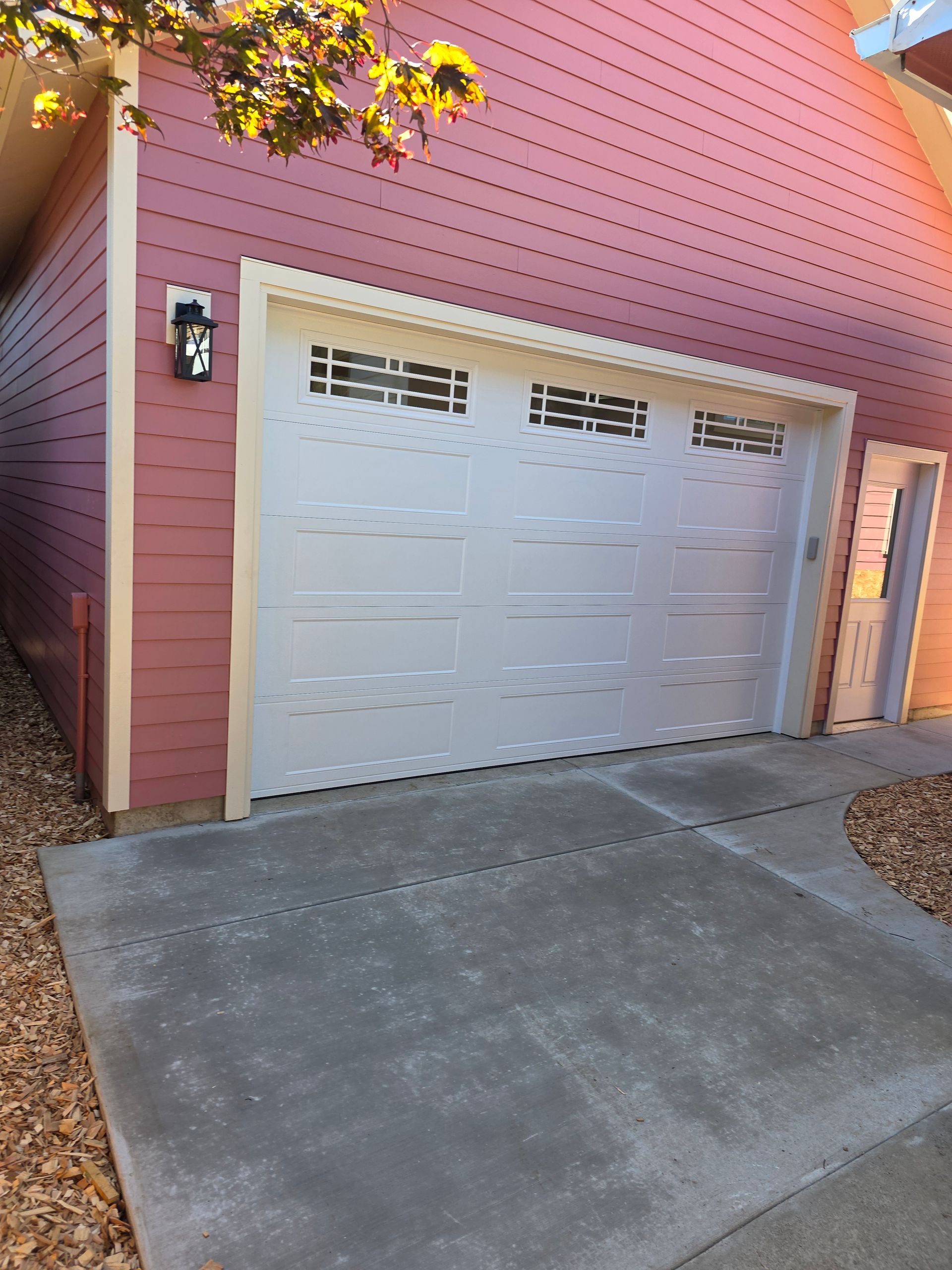 Red double garage door on a pink house with a concrete driveway and autumn leaves overhead