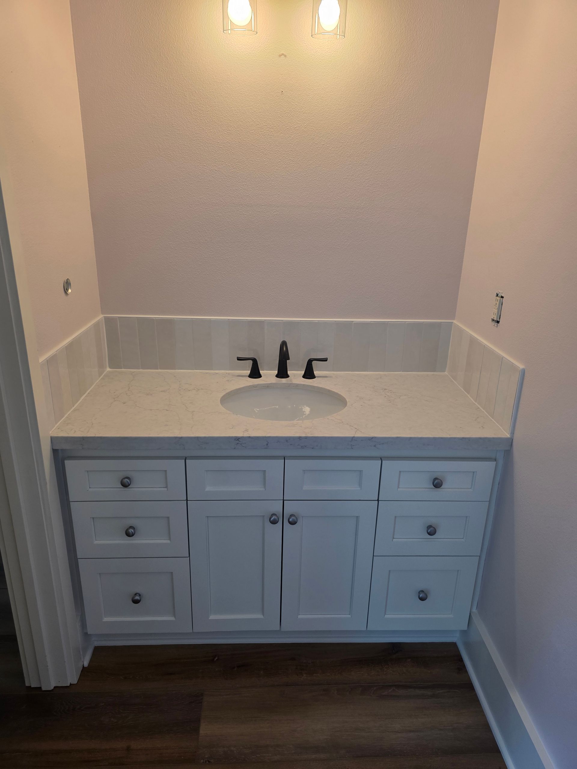 Small bathroom vanity with white cabinet, sink, mirror, and wall light over dark wood floor.