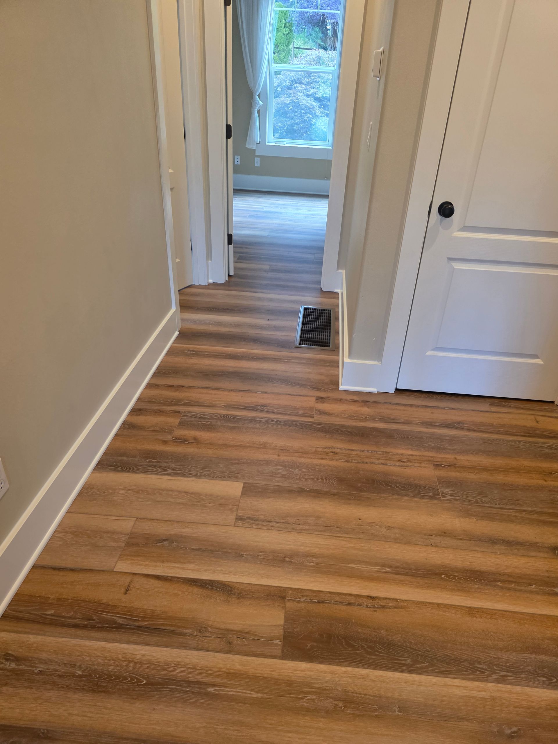 Empty hallway with wood-look flooring and white trim, leading to a doorway and a window at the end.