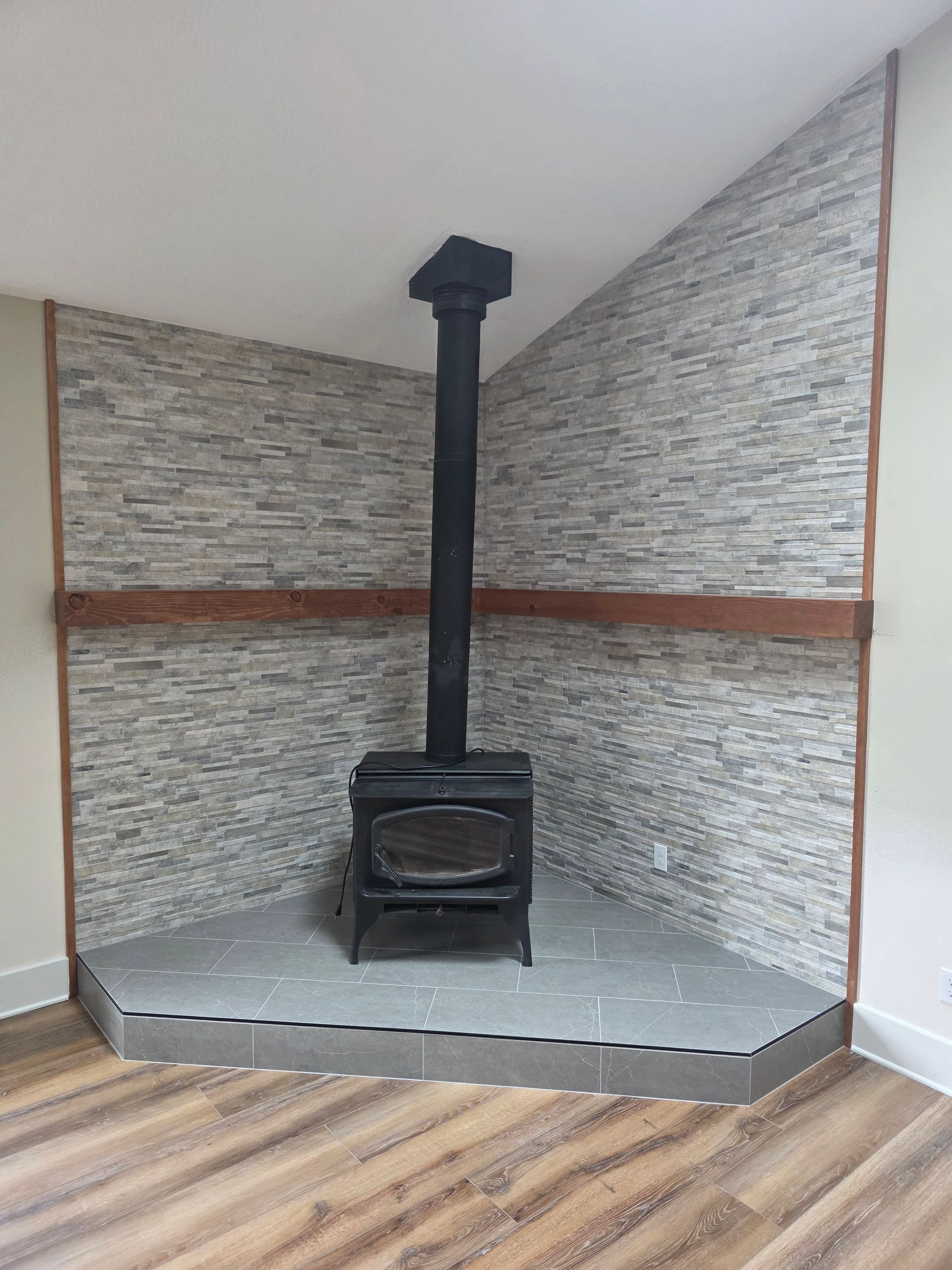 Black wood stove with chimney in a small corner alcove, against gray stone walls and a wood floor.